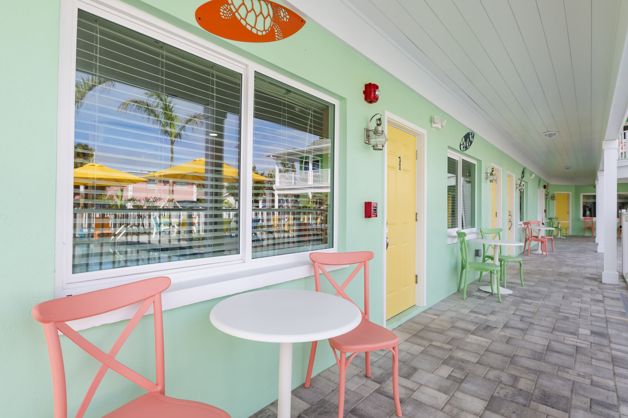 An outdoor walkway of a mint green building with white trim and a grey stone-tiled floor. In the foreground, a small white round table is flanked by two coral-colored chairs. A large window with white blinds reflects palm trees and yellow umbrellas. Further down the corridor, a yellow door and additional seating areas are visible under a white-paneled ceiling.