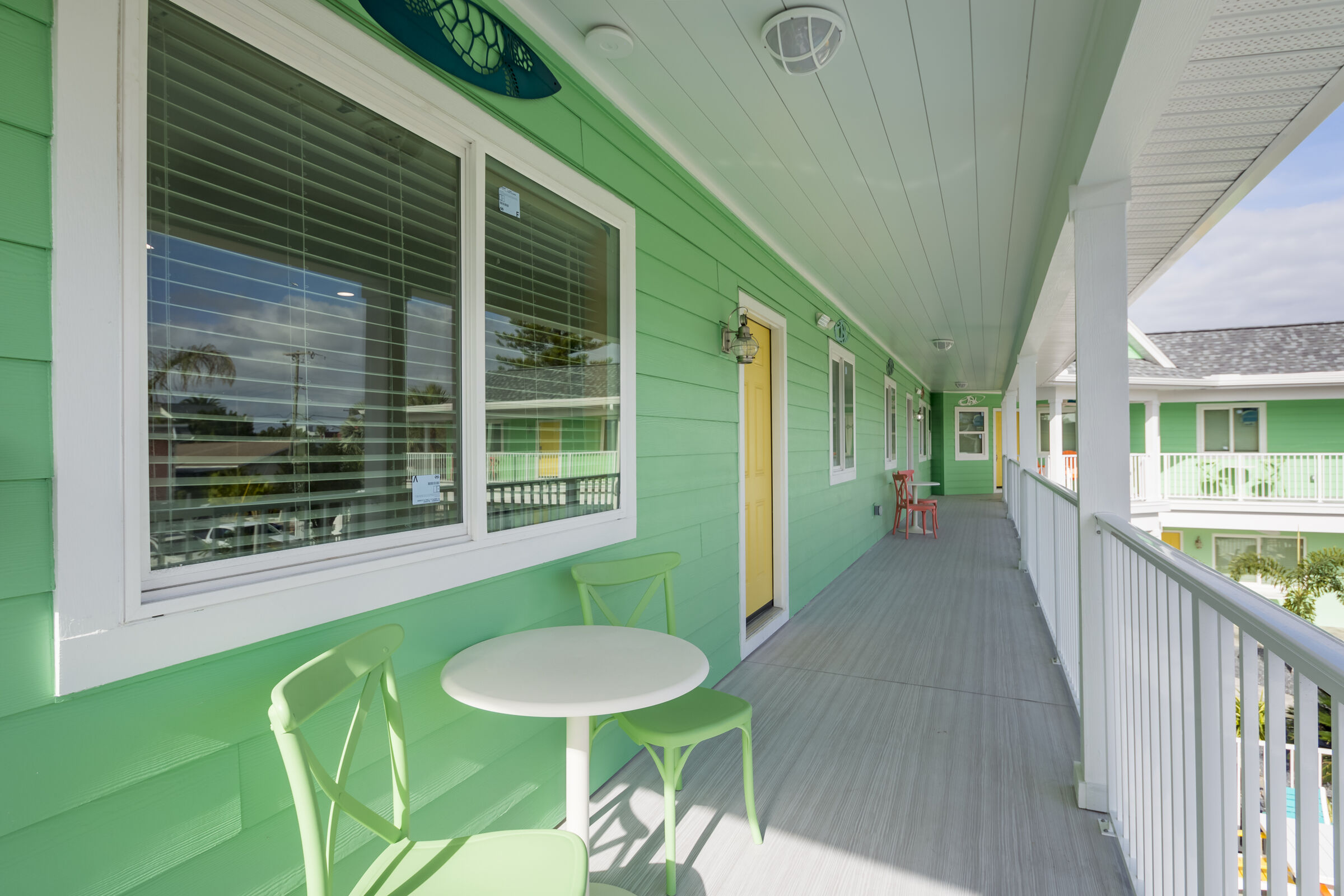 An exterior view of a mint green building with a white-railed walkway. A small white café table and two lime green chairs sit outside a window, with a yellow door visible further down the path.