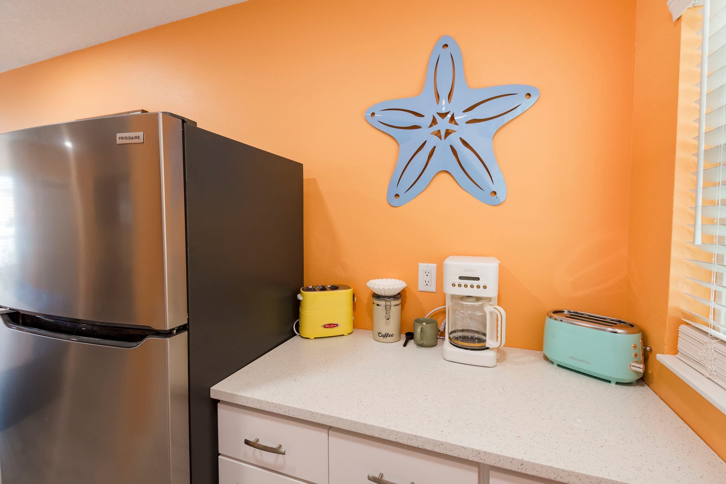A white countertop area against an orange wall featuring a large, light blue decorative starfish. The counter holds a small yellow appliance, a white coffee maker, and a mint green toaster. To the left, a stainless steel refrigerator is partially visible, and to the right, a window with white blinds lets in bright light.
