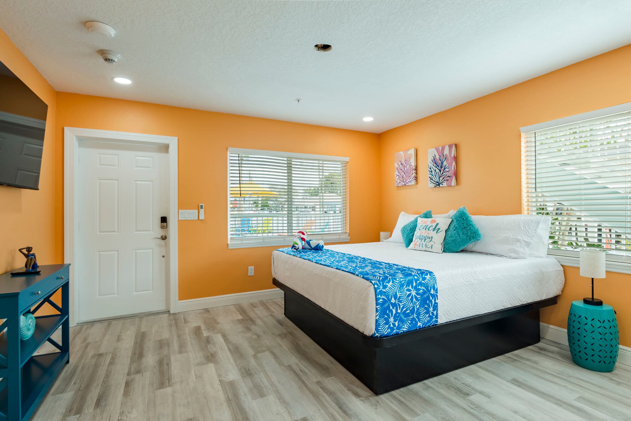 A spacious studio apartment with vibrant orange walls and light wood-look flooring. A large bed with white linens and a blue floral runner sits in the center of the room, flanked by windows with white blinds. To the left, there is a white entry door and a blue console table, while the right side of the room features a teal side table with a small lamp.