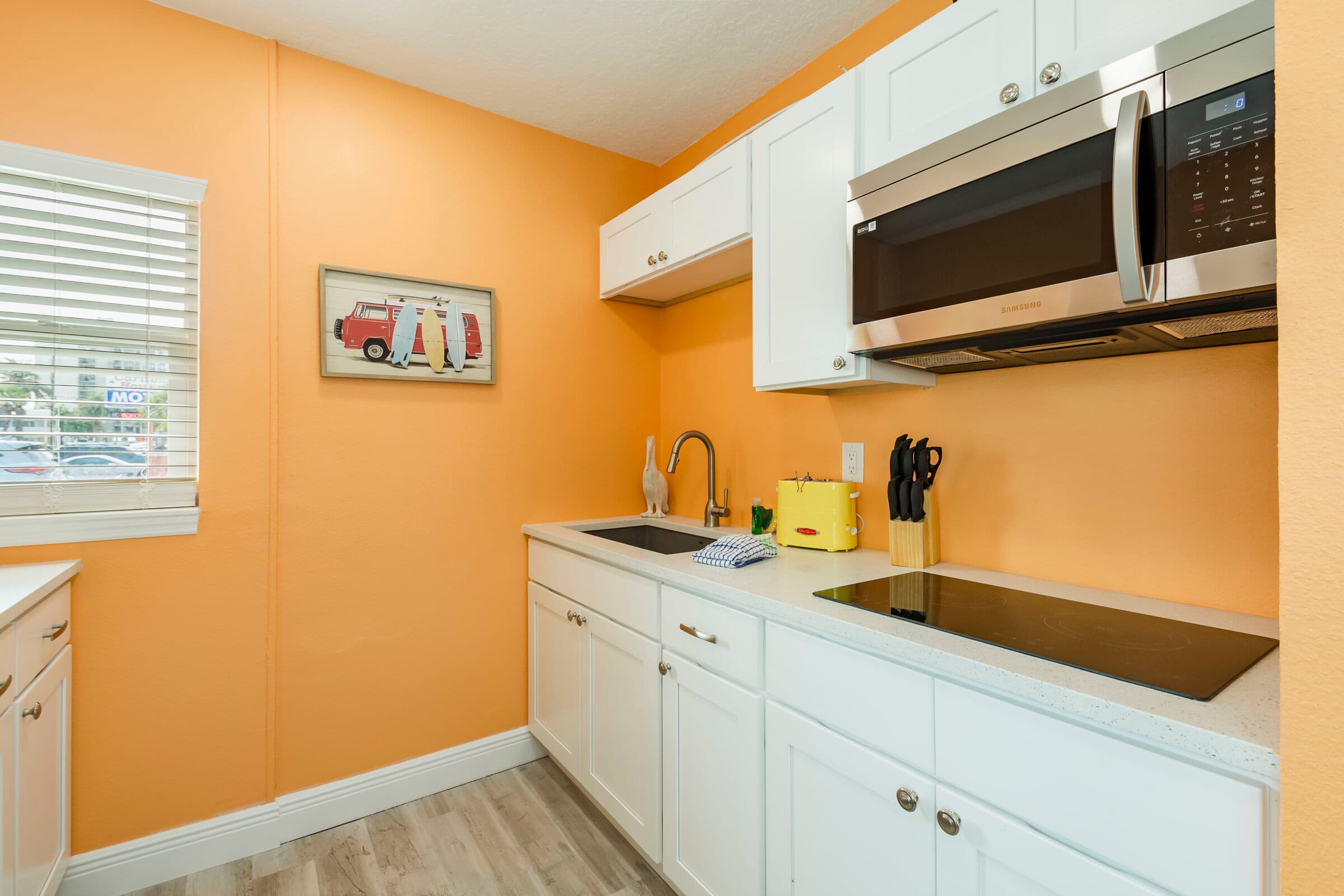 A modern kitchenette with bright orange walls and white cabinetry. The countertop features a built-in cooktop, a stainless steel sink with a high-arc faucet, and a small yellow toaster. A black microwave is mounted above the counter, and a small framed artwork depicting surfboards hangs on the wall next to a window with white blinds.
