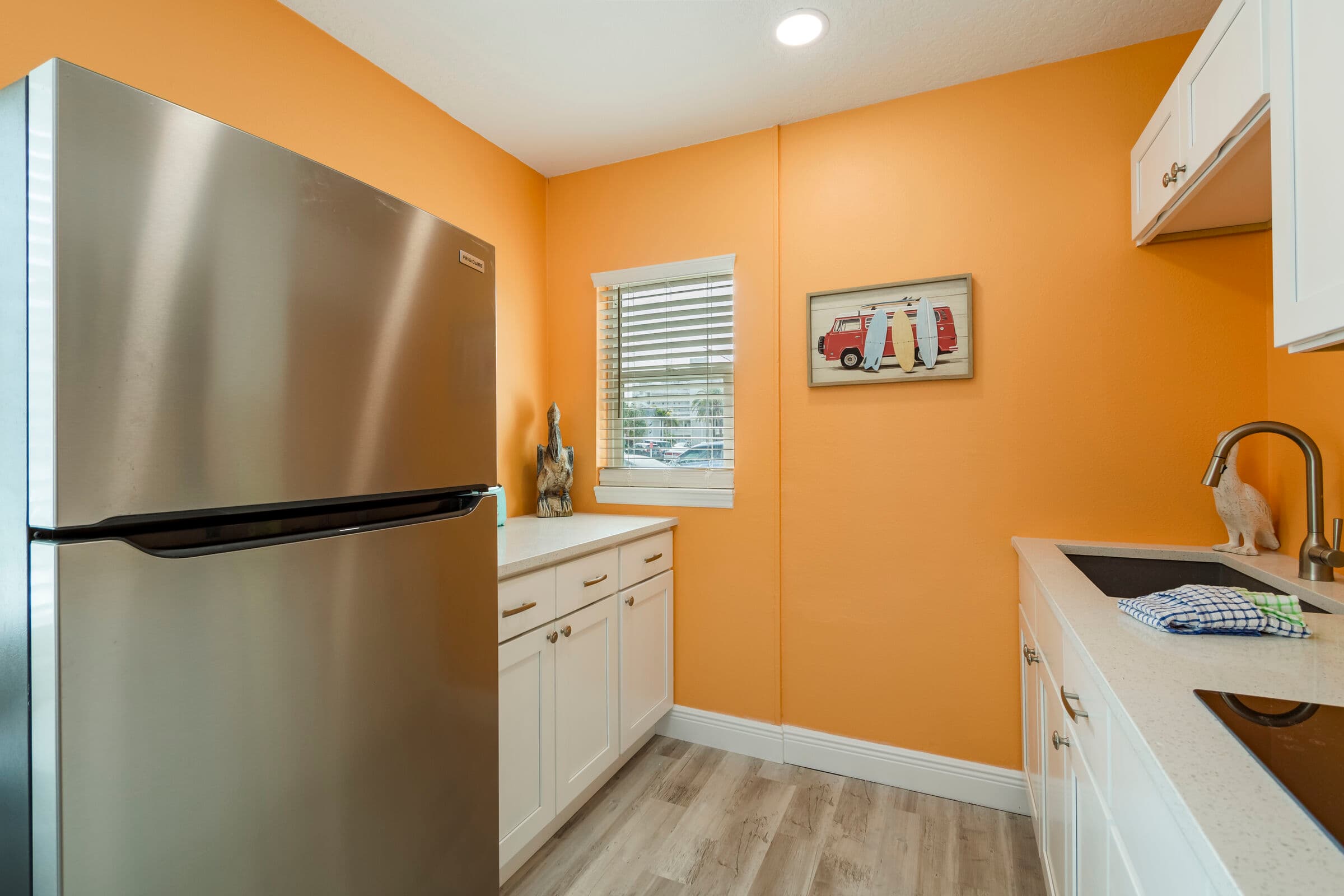 A compact kitchenette featuring bright orange walls and light wood flooring. A large stainless steel refrigerator stands on the left, while white cabinets and a white countertop run along both sides of the room. The space includes a sink with a gooseneck faucet and a small window with white blinds that lets in natural light.