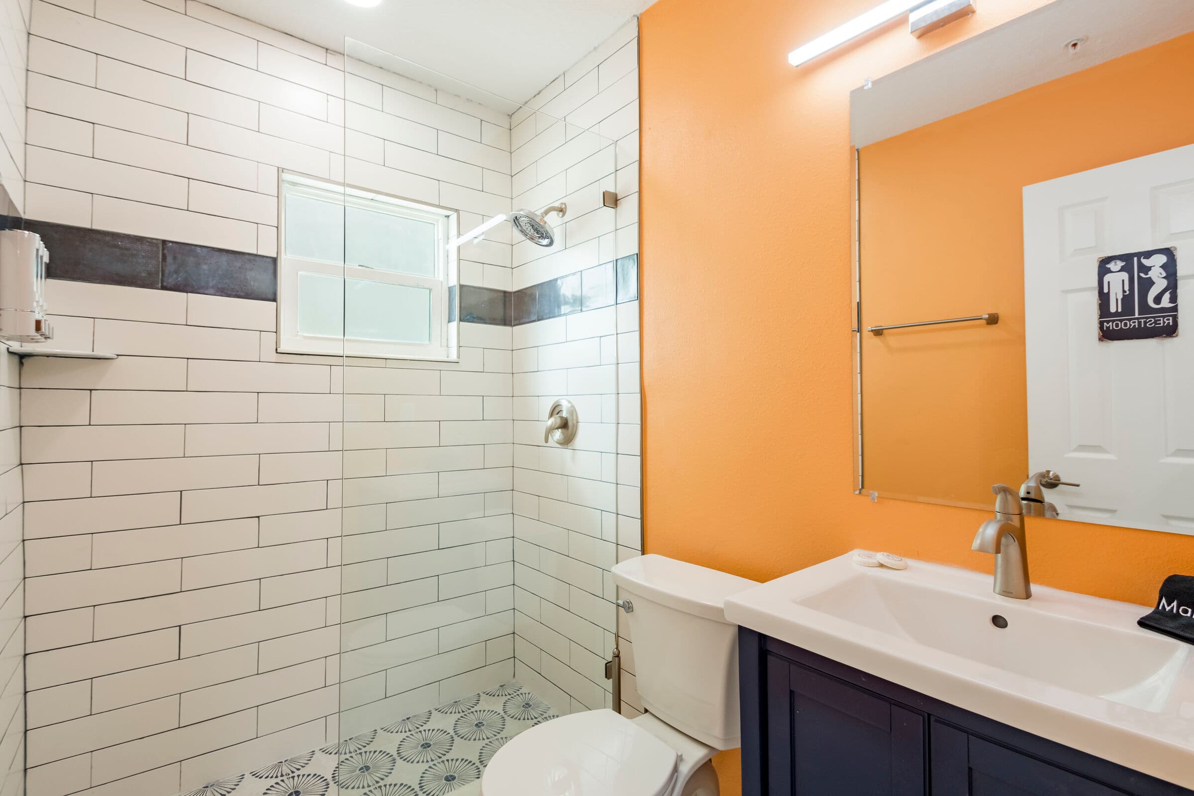 A modern bathroom featuring bright orange walls and white subway tile in the shower area. A navy blue vanity with a white ceramic sink and a chrome faucet is positioned beneath a large frameless mirror. The walk-in shower includes a glass partition, a gray decorative tile accent strip, and patterned floor tiles.