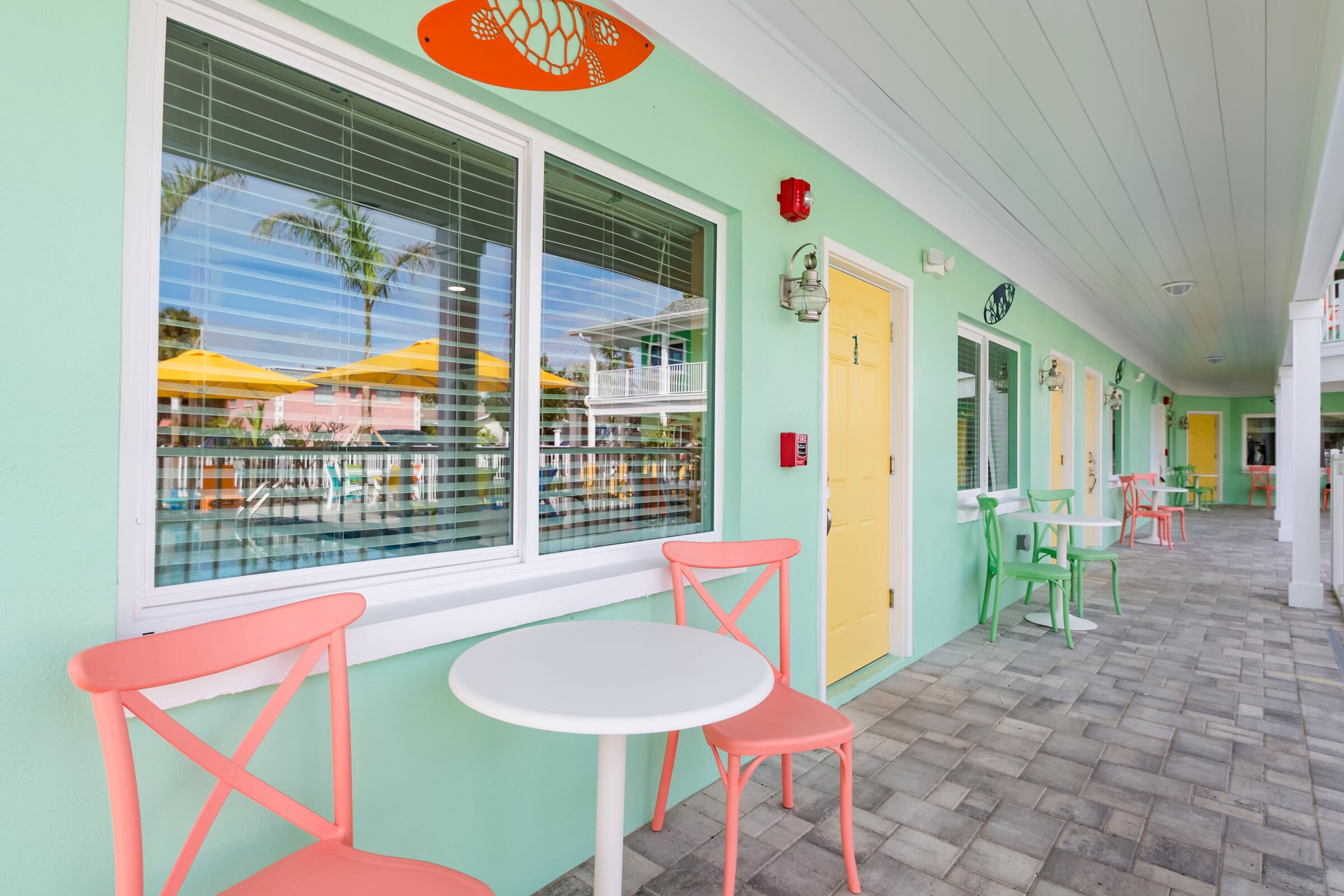 An outdoor walkway of a mint green building with white trim and a grey stone-tiled floor. In the foreground, a small white round table is flanked by two coral-colored chairs. A large window with white blinds reflects palm trees and yellow umbrellas. Further down the corridor, a yellow door and additional seating areas are visible under a white-paneled ceiling.