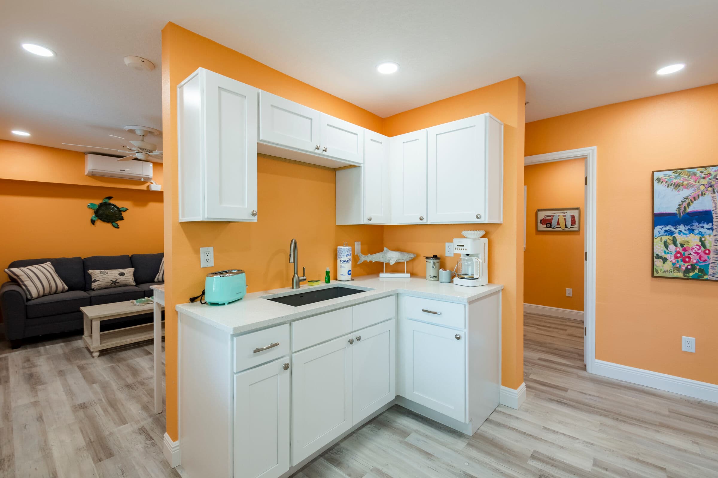 A modern kitchenette featuring white cabinetry and a white countertop against vibrant orange walls. The space includes a built-in black cooktop, a stainless steel sink with a high-arc faucet, and a black microwave mounted above the counter. A small yellow toaster sits on the counter, and a piece of beach-themed artwork hangs on the wall near a window with white blinds.