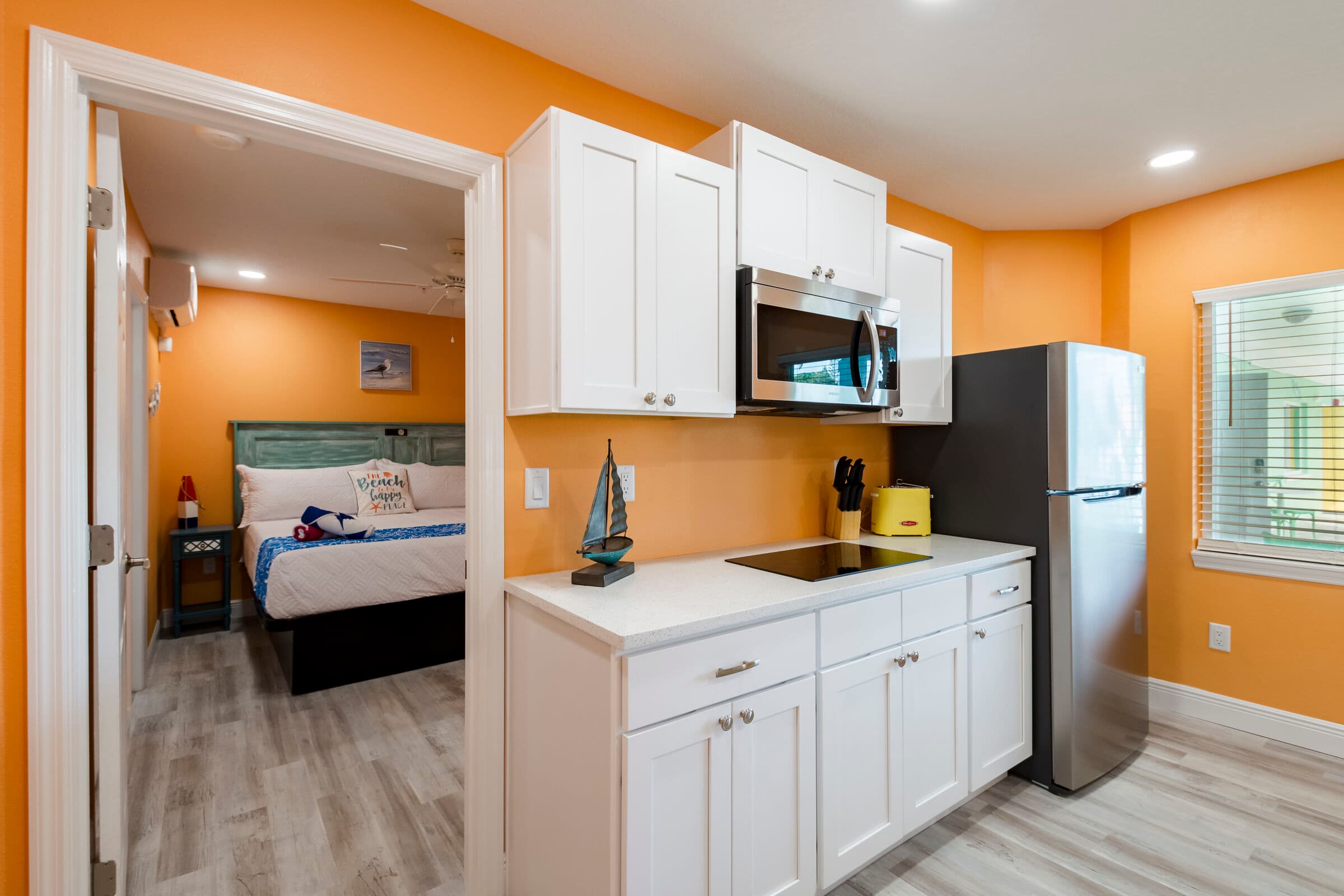 A modern kitchenette featuring white cabinetry and a white countertop against vibrant orange walls. The space includes a built-in black cooktop, a stainless steel refrigerator, and a stainless steel microwave mounted above the counter. To the left, an open doorway reveals a bedroom with a large bed and light wood-look flooring.