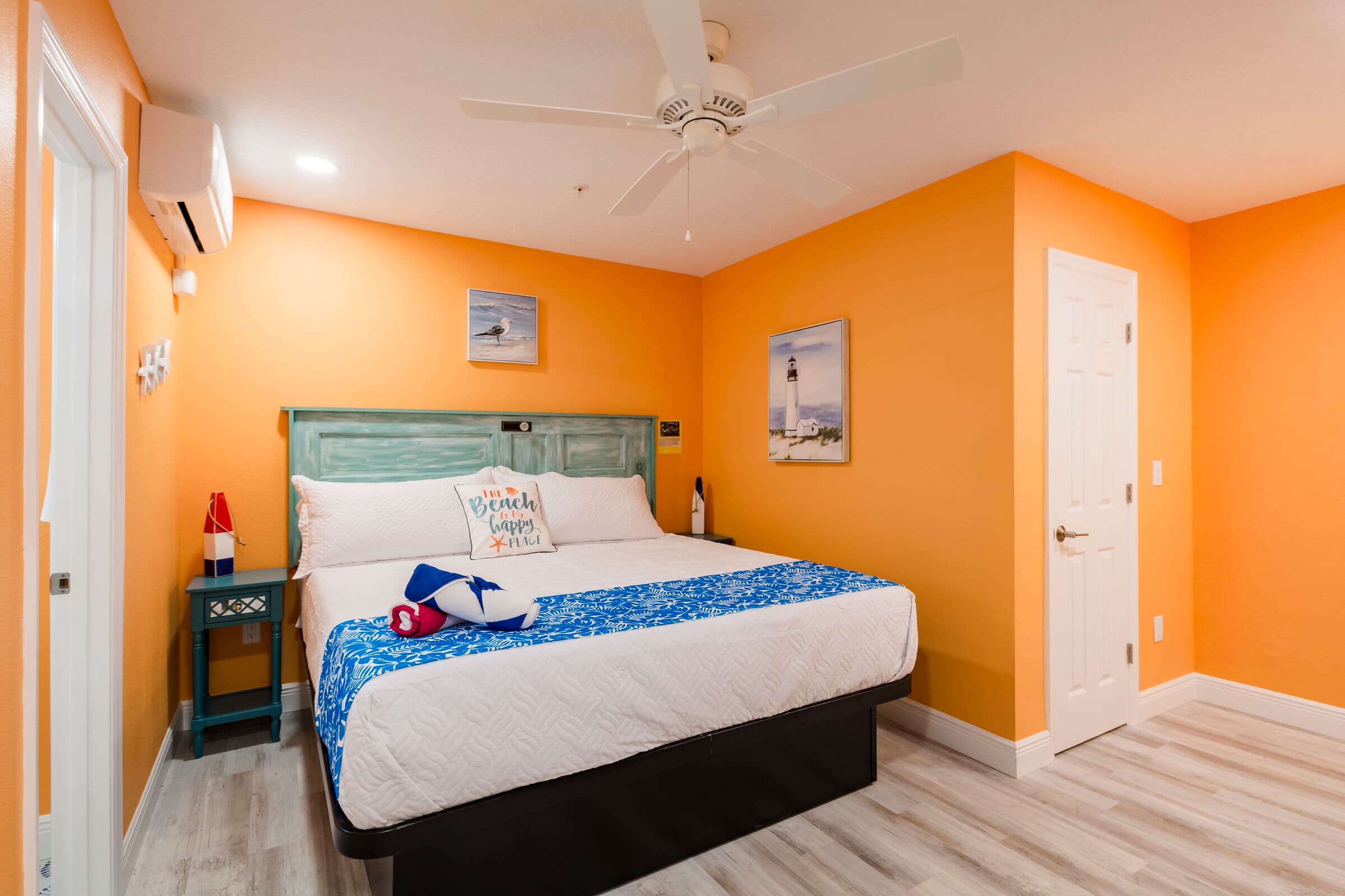 A cozy bedroom with bright orange walls and light wood-look flooring features a large bed with a teal, rustic wood headboard. The bed is dressed in white linens, accented by a blue floral runner and matching pillows. To the left of the bed is a small blue nightstand with a decorative red and white buoy lamp, and beach-themed artwork hangs on the walls.