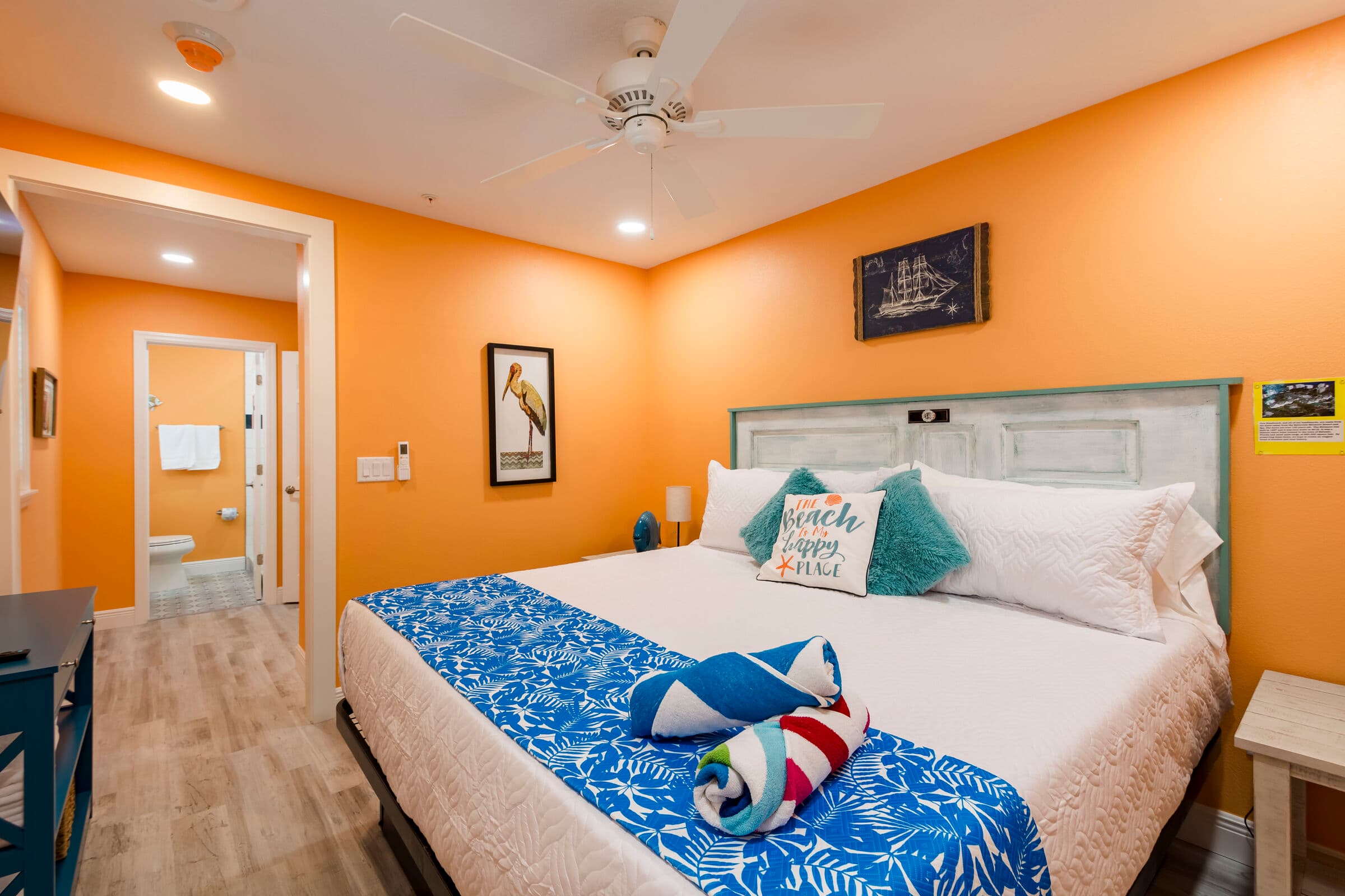 A cozy studio apartment with vibrant orange walls and light wood-look flooring. In the foreground, a large bed is made with white linens, a blue-and-white floral runner, and turquoise accent pillows. To the left, a dark blue console table sits against the wall, and an open doorway leads to a bright white-tiled bathroom in the background.
