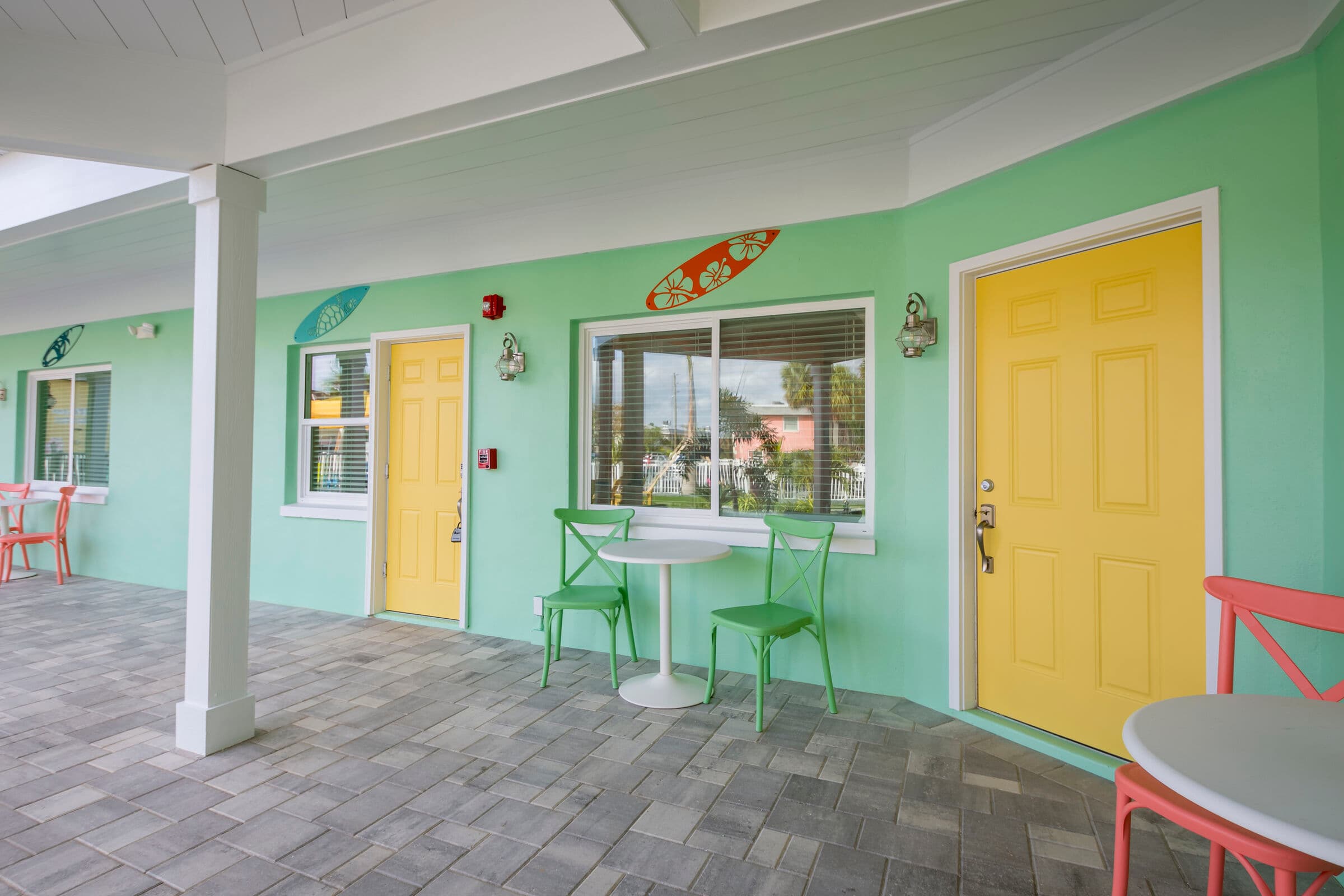 An outdoor corridor of a mint green building with yellow doors and stone tile flooring. The scene features multiple small seating areas, each with a white round table and two colorful chairs in coral or green. Decorative surfboards are mounted above the windows, and the white-paneled ceiling is supported by white pillars.