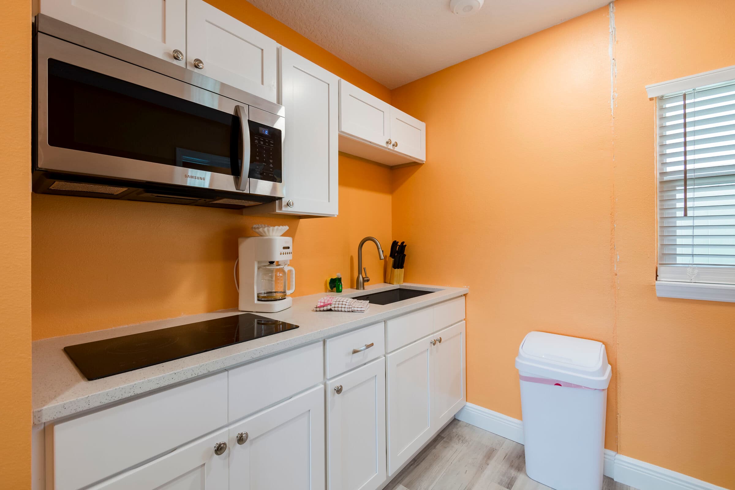 A bright kitchenette with vibrant orange walls and white cabinetry. The setup features a white countertop with a built-in black cooktop, a stainless steel sink with a high-arc faucet, and a large stainless steel microwave mounted above. A white coffee maker sits on the counter, and a white trash can is placed on the light wood-look flooring.