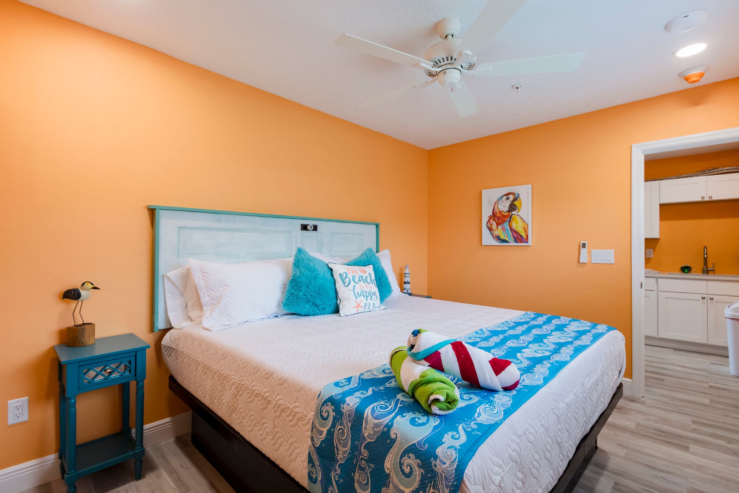 A cozy bedroom with vibrant orange walls and light wood-look flooring features a large bed with a teal, rustic wood headboard. The bed is dressed in white linens, accented by a blue-and-white floral runner and matching pillows. To the left is a small teal nightstand with a decorative lamp, and a white doorway on the right leads to a kitchenette area.