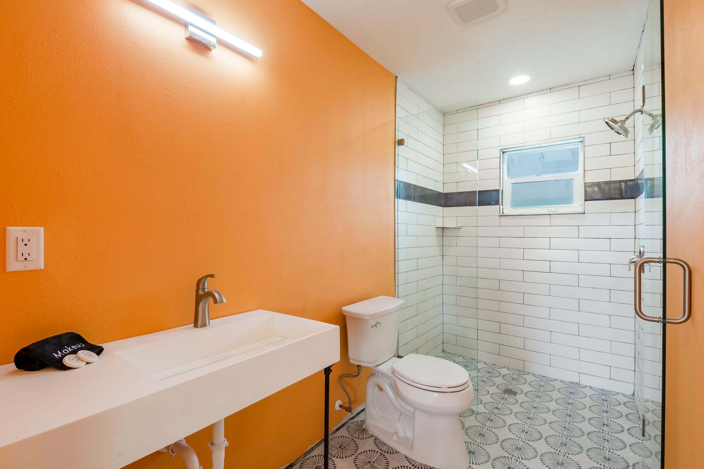 A modern bathroom with orange walls featuring a white trough-style sink and a white toilet. The space includes a glass-enclosed walk-in shower with white subway tile, a decorative blue tile accent strip, and patterned floor tiles.