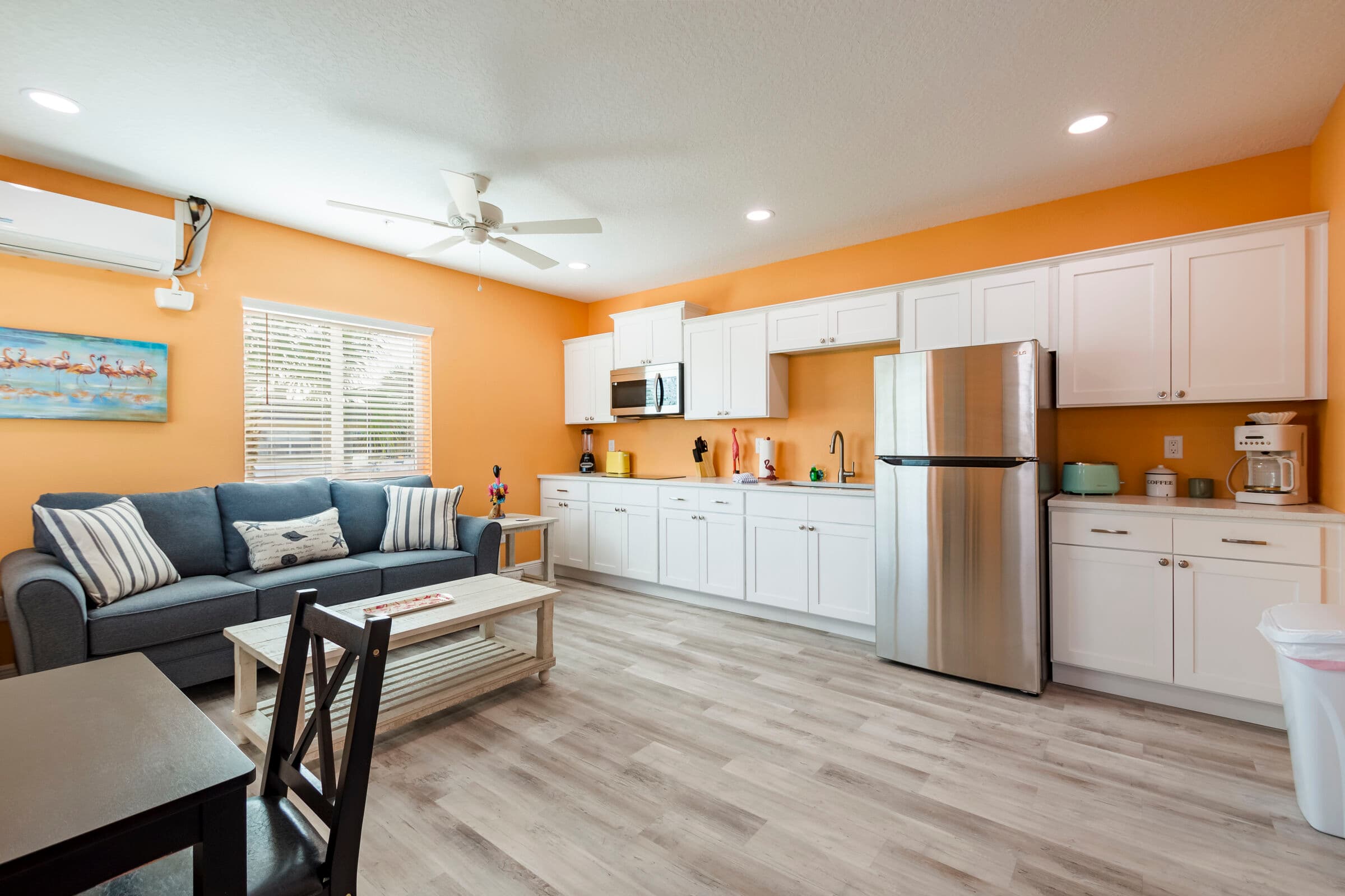 A spacious, open-concept living and dining area featuring orange walls and light wood-style flooring. The space includes a grey sofa with striped pillows, a white coffee table, and a full wall of white kitchen cabinets with stainless steel appliances, including a large refrigerator and a microwave.
