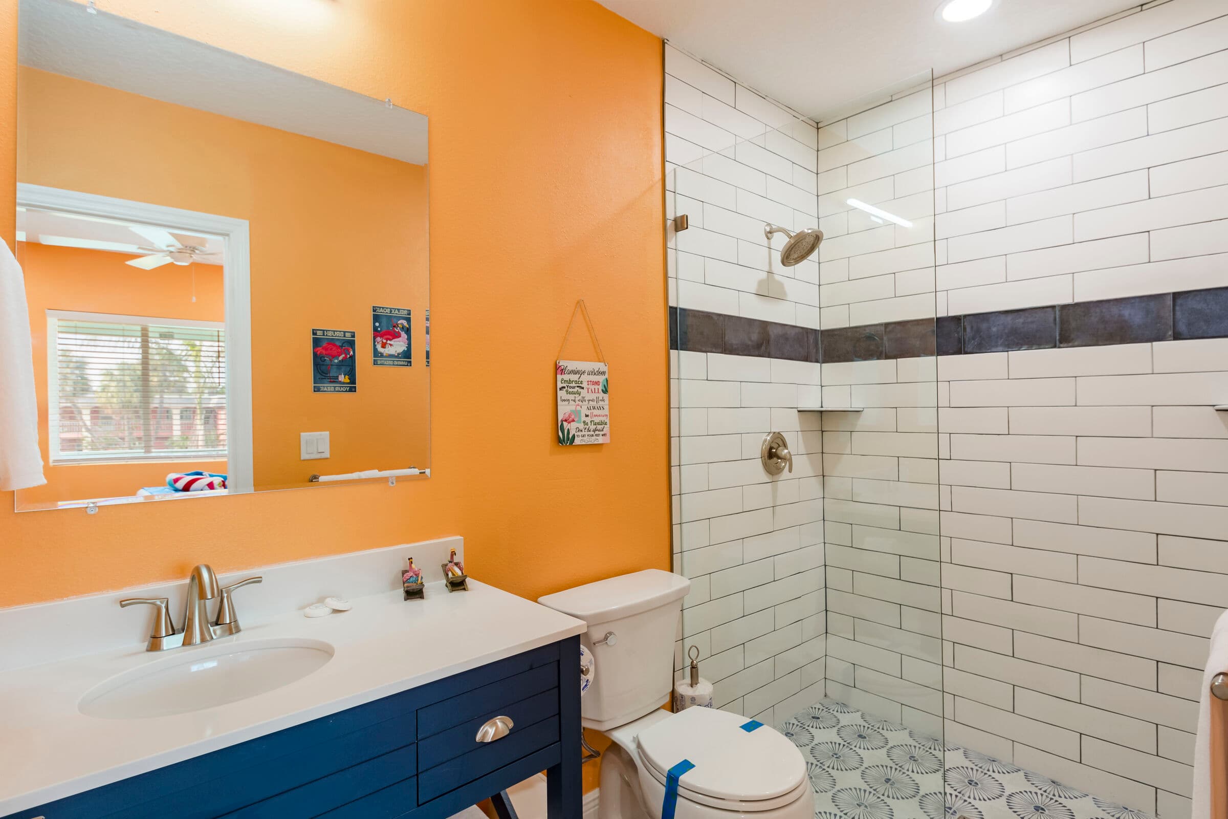 A modern bathroom with orange walls and a dark blue vanity featuring a white countertop and silver faucet. The space includes a white toilet and a glass-enclosed walk-in shower with white subway tiles, a decorative dark grey accent strip, and patterned grey floor tiles.