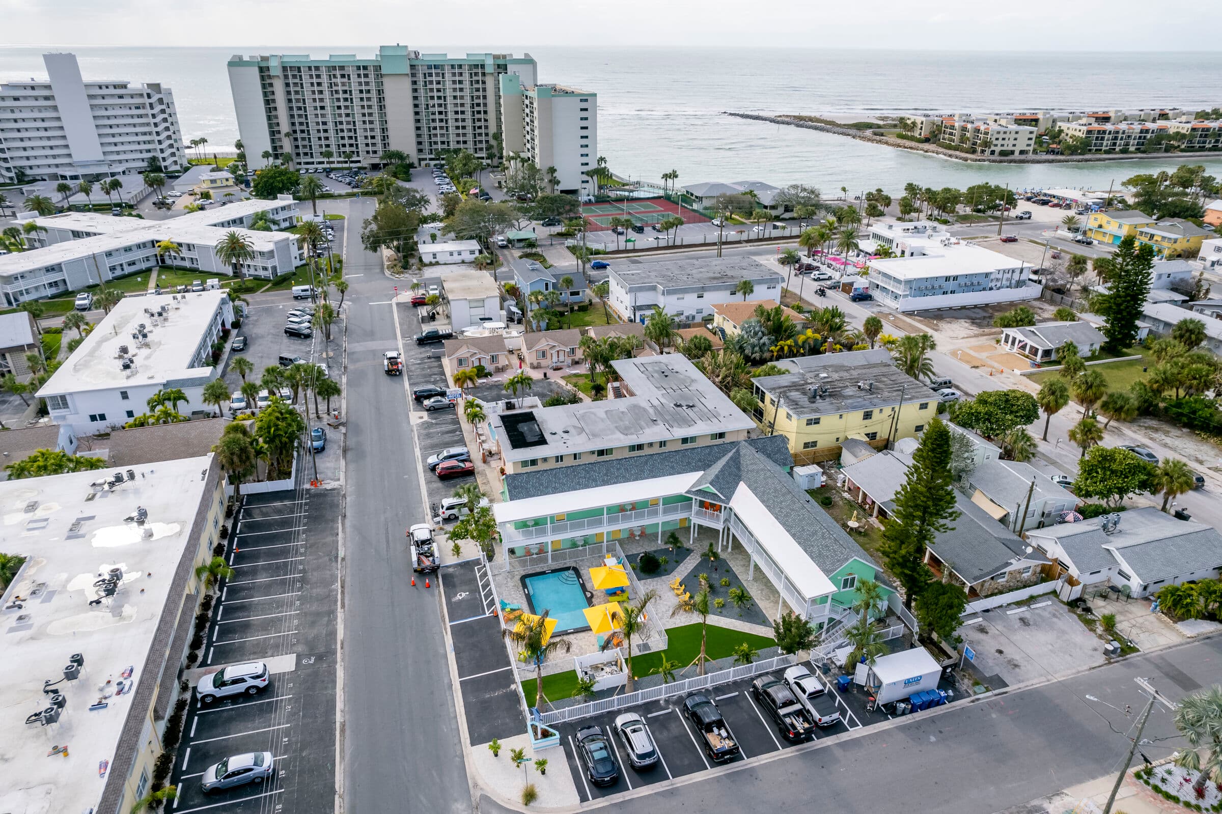 An aerial view of a coastal neighborhood featuring a two-story L-shaped motel with a swimming pool and yellow umbrellas. The surrounding area includes residential streets, parking lots, and large hotel buildings overlooking a nearby bay and ocean in the background.