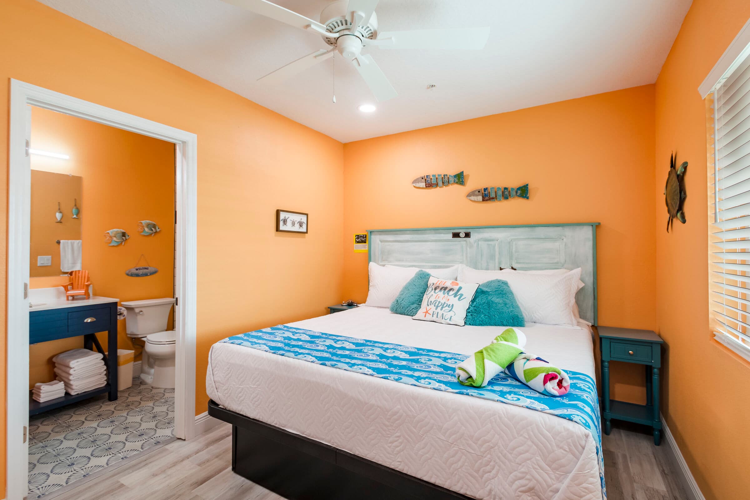 A bedroom with vibrant orange walls and light wood-style flooring, featuring a large bed with a white headboard and a blue patterned bed runner. A doorway on the left leads into a bathroom with a dark blue vanity, and several beach-themed art pieces, including metal fish, decorate the walls.