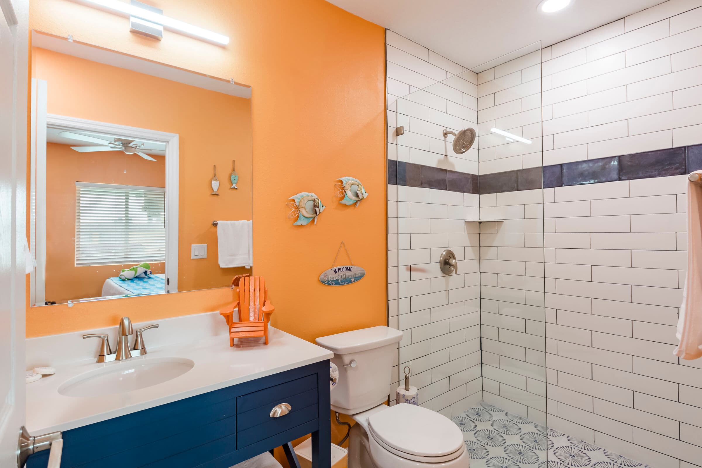 A modern bathroom with vibrant orange walls and a dark blue vanity with a white countertop. The space includes a white toilet and a glass-enclosed walk-in shower featuring white subway tiles with a dark accent strip and patterned grey floor tiles.
