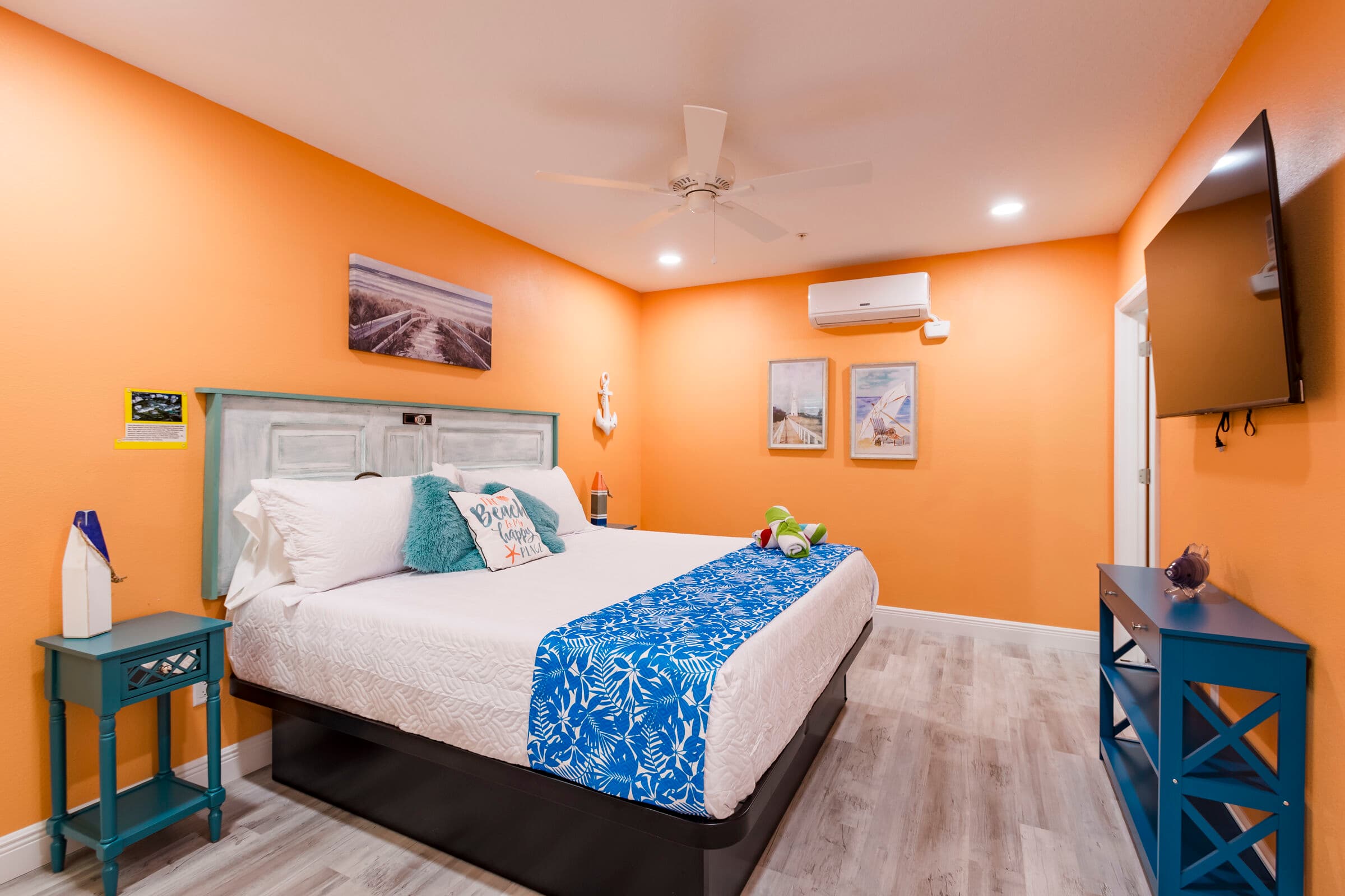 A cozy bedroom with vibrant orange walls and light wood-style flooring, featuring a large bed with a white headboard and a blue patterned bed runner. A tall wall-mounted TV hangs on the right wall above a blue console table, while beach-themed artwork and a white ceiling fan complete the coastal decor.