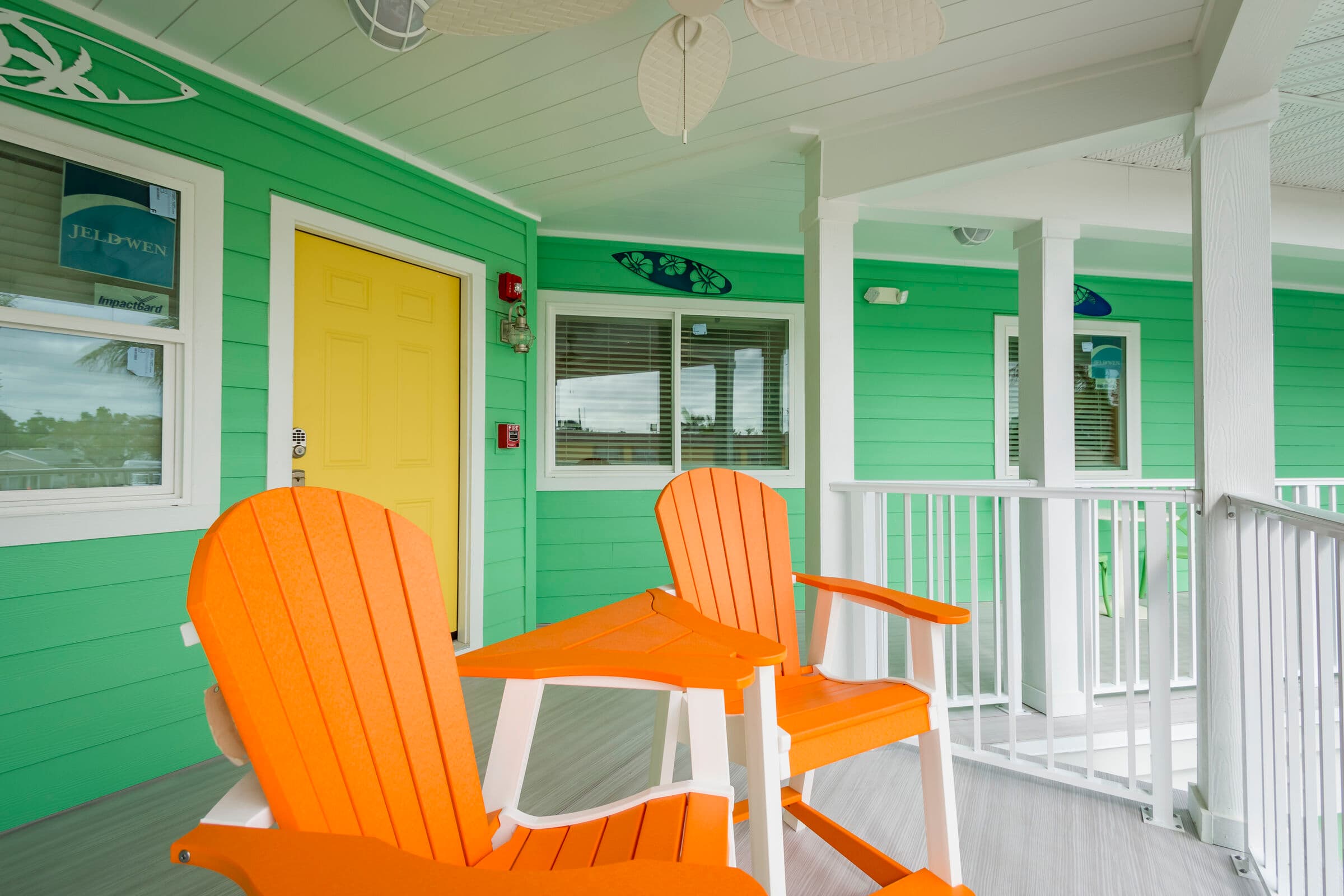 Two bright orange Adirondack chairs and a small white side table sit on a white-railed balcony outside a mint green building. A yellow door and a window with white trim are visible in the background, along with a decorative surfboard mounted on the wall.
