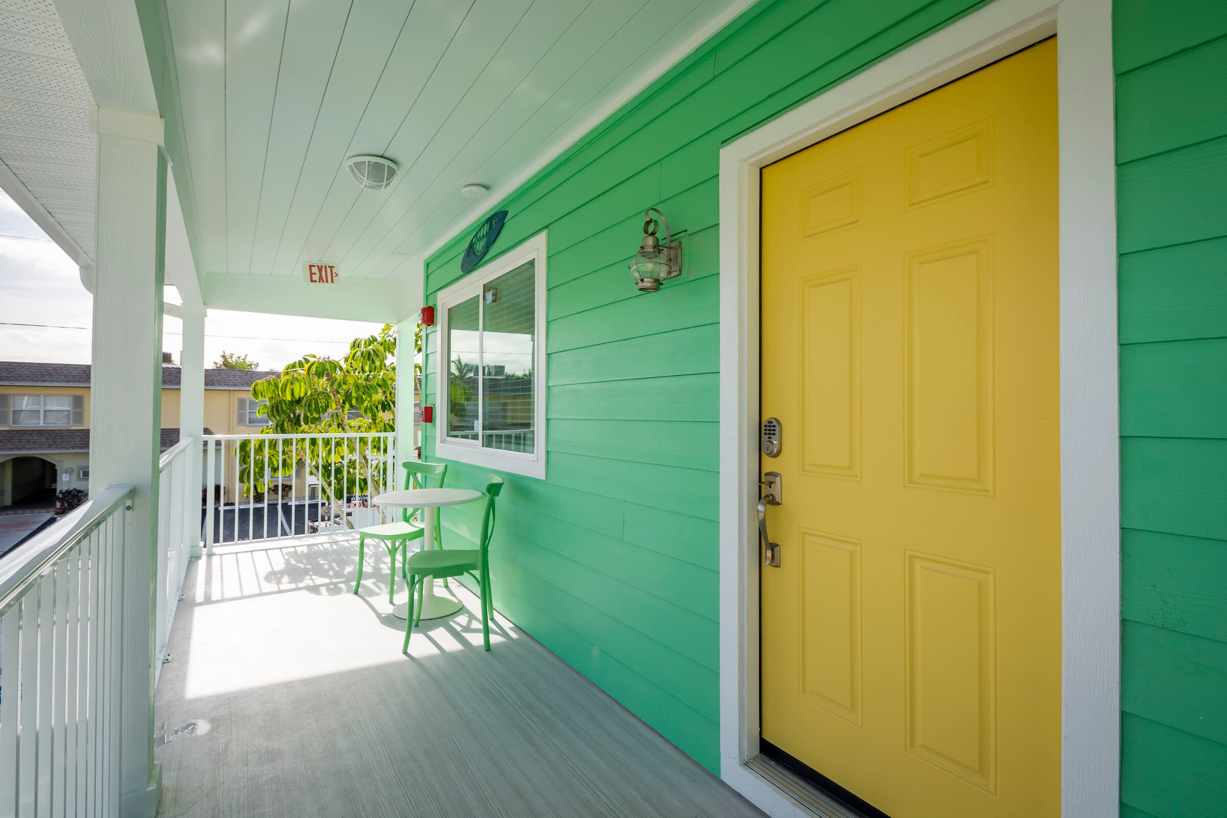 A bright green balcony featuring a vibrant yellow door, white railings, and a small bistro set with two green chairs and a white table.