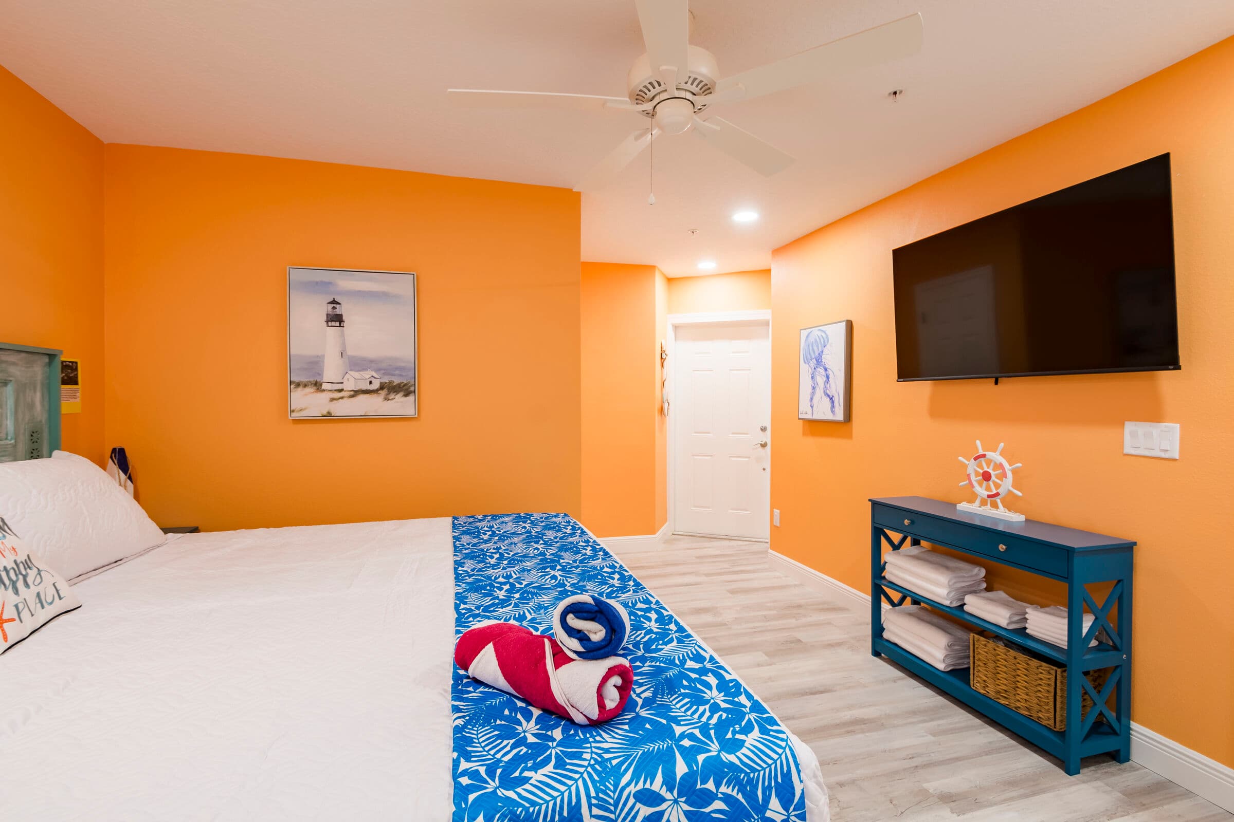 A hotel bedroom with vibrant orange walls and light grey flooring, featuring a bed with a blue and white patterned runner and a navy blue console table beneath a wall-mounted television.