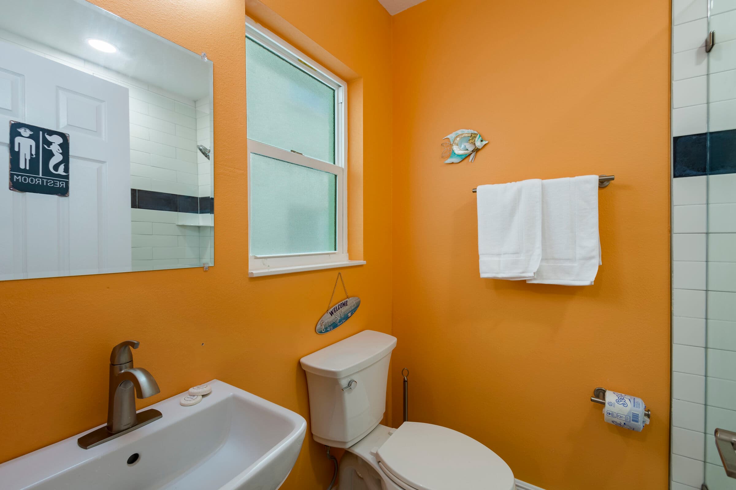 A vibrant bathroom featuring warm orange walls and patterned grey floor tiles. It includes a white pedestal sink, a standard toilet, and a walk-in shower lined with white subway tiles and a navy blue accent stripe.