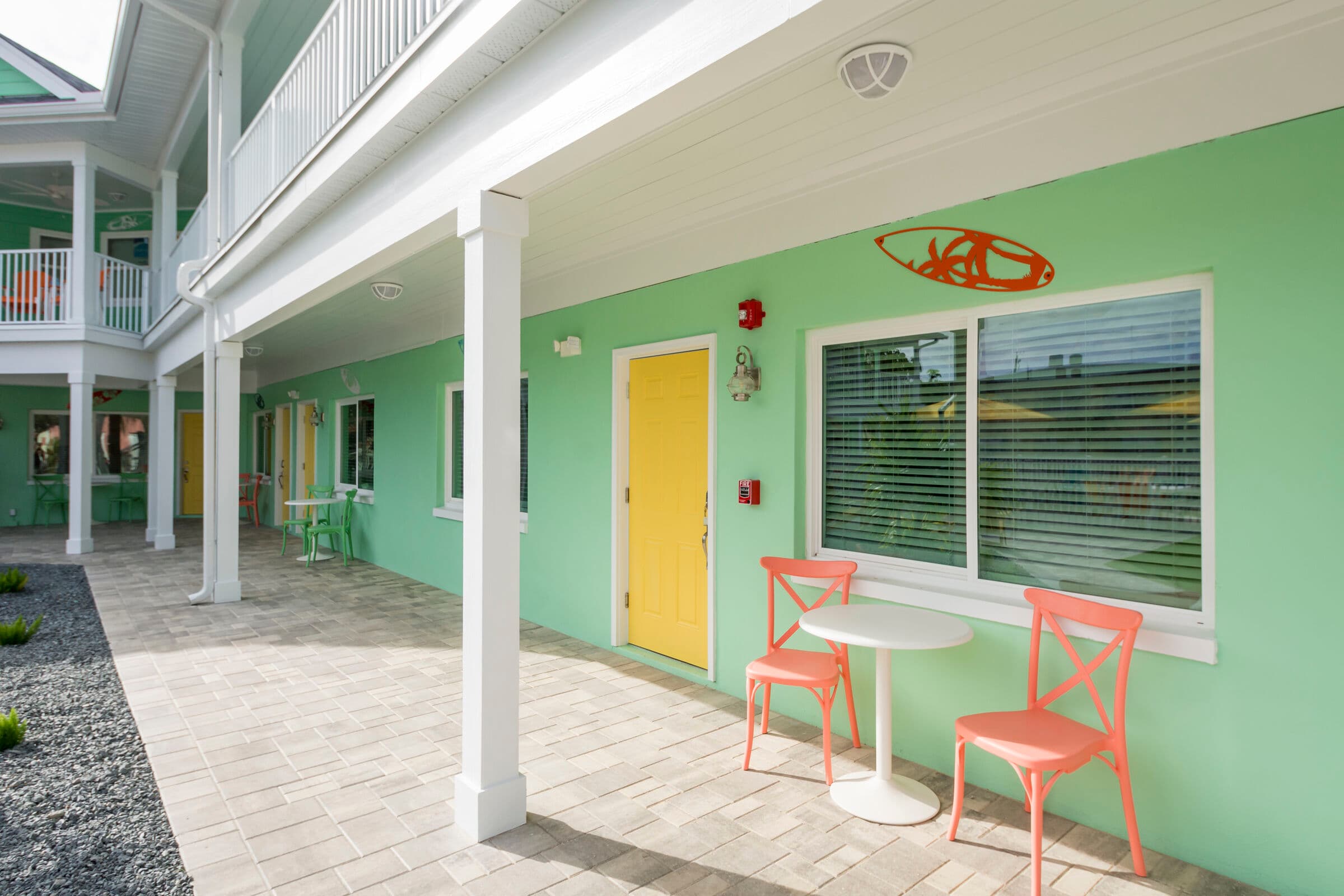 An outdoor patio walkway of a mint green building featuring a yellow door and white pillars. A small white bistro table with two coral-colored chairs sits in front of a window with white blinds. A decorative orange surfboard is mounted on the wall above the window, and the ground is paved with grey stone tiles.