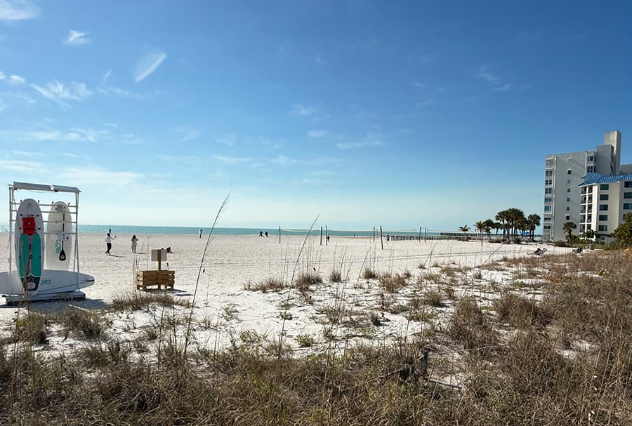 A sandy beach with distant figures walking, a lifeguard stand, and a high-rise building under a clear blue sky.