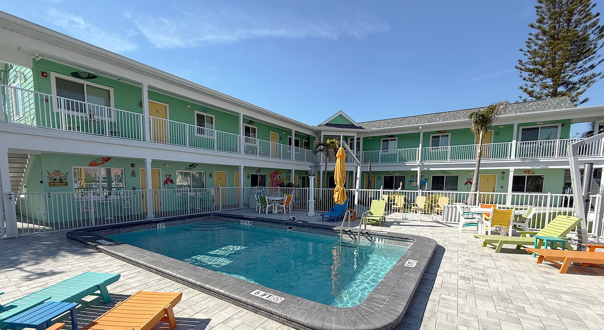 A bright courtyard featuring a swimming pool surrounded by colorful lounge chairs and green hotel rooms under a clear blue sky.