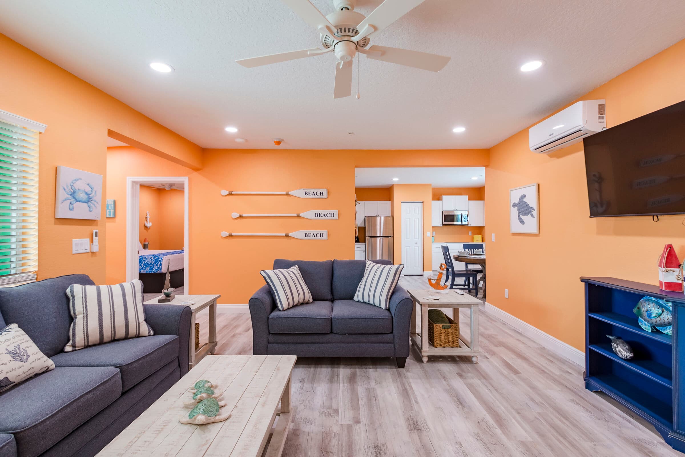 A spacious living area with orange walls and light wood-plank flooring, featuring two grey sofas with striped pillows, a white coffee table, and a wall-mounted TV over a blue console. The background shows three decorative oars mounted on the wall and a glimpse into a bedroom and a dining area.