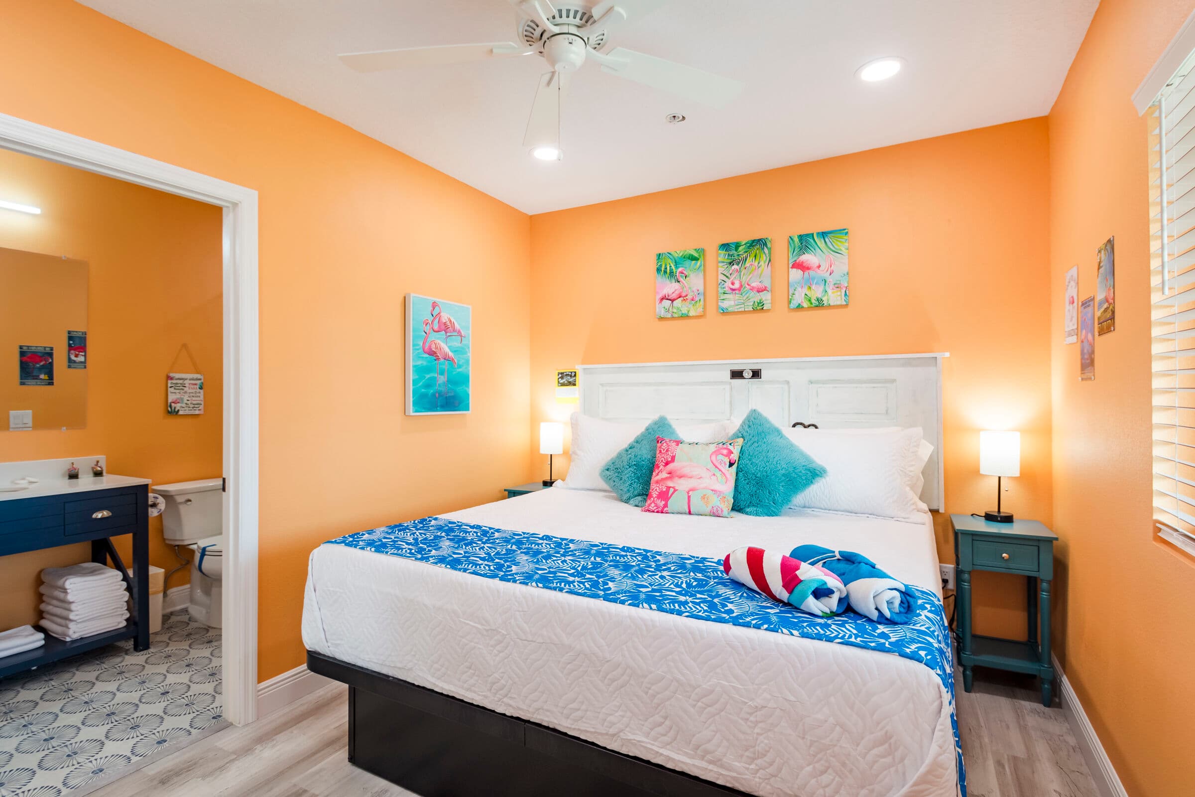 A cozy bedroom with vibrant orange walls and light wood-style flooring, featuring a large bed with a white headboard and a blue patterned bed runner. A doorway on the left provides a view into a bathroom with a dark blue vanity, while several beach-themed art pieces decorate the walls.