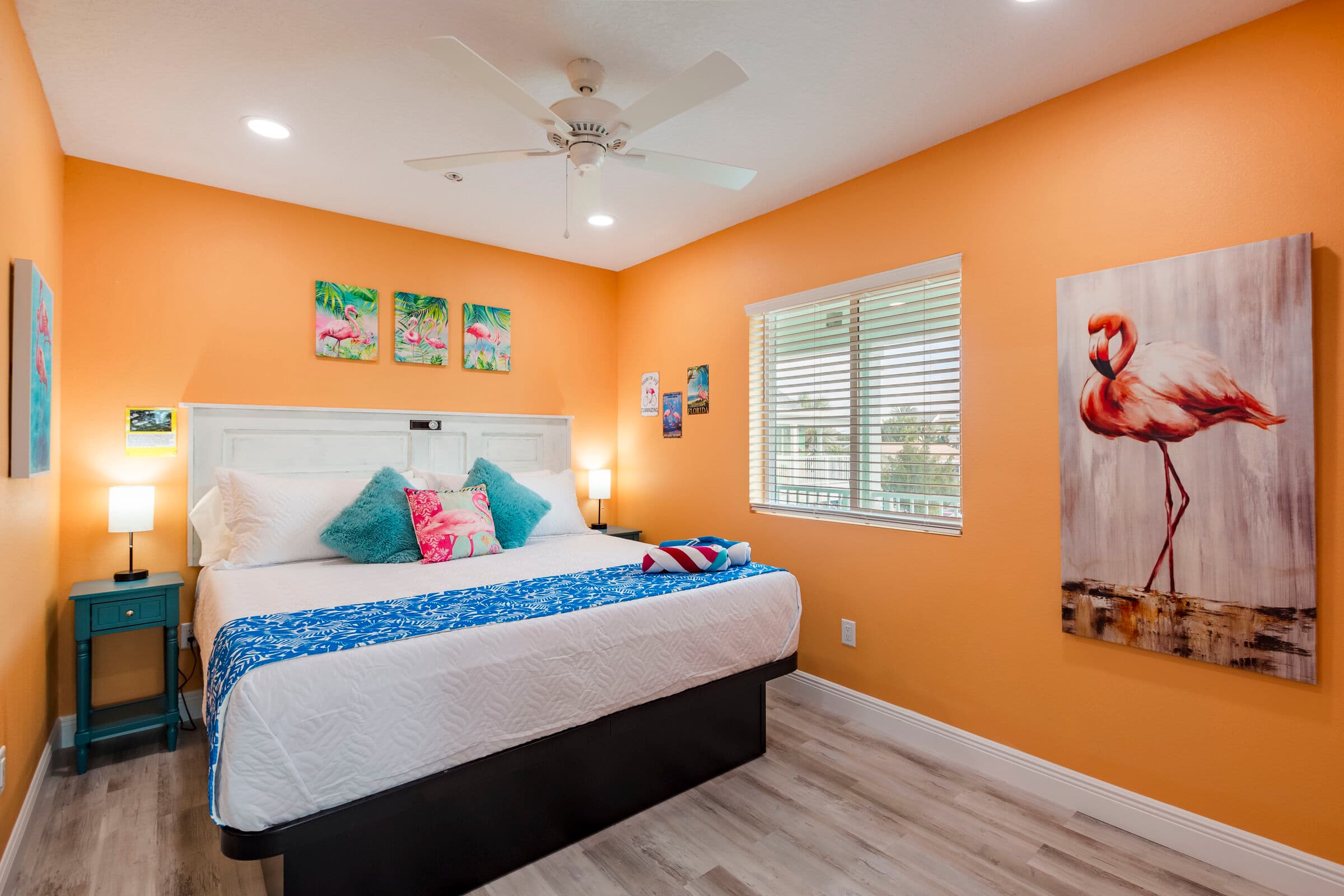 A bright bedroom with vibrant orange walls and light wood-style flooring, featuring a large bed with a white headboard and a blue patterned bed runner. A tall canvas print of a pink flamingo hangs on the right wall, and three smaller beach-themed artworks are displayed above the headboard.