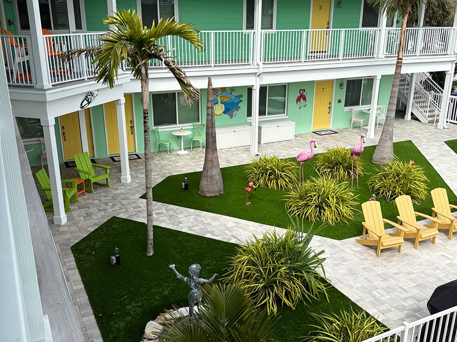 A colorful courtyard with tropical plants, palm trees, and vibrant doors surrounds a lawn dotted with flamingos and seating areas.