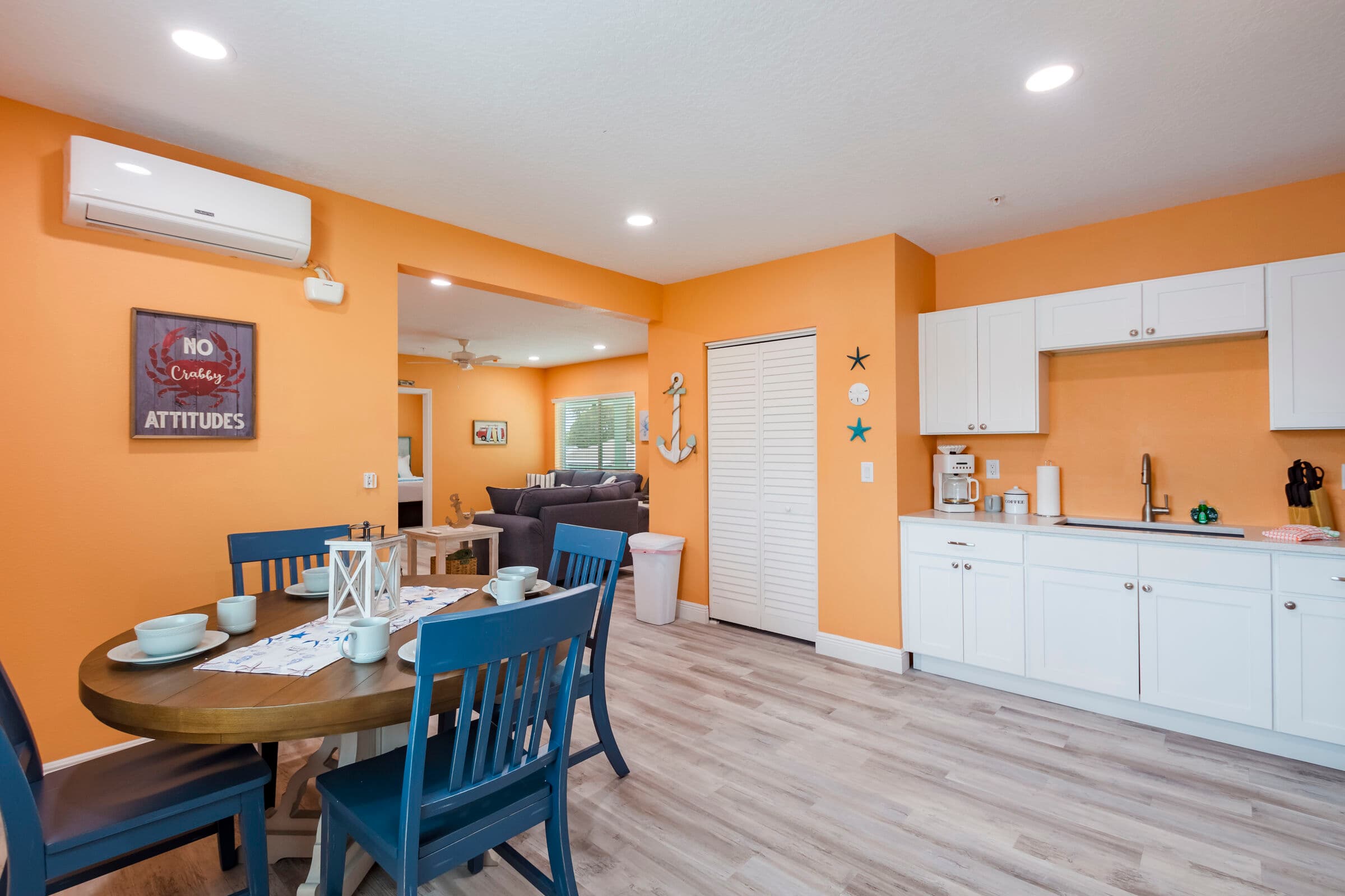 A spacious, open-concept living area and kitchen with orange walls and light wood-style flooring. The room features a grey sofa with striped pillows, a white coffee table, a small black dining set, and a full wall of white kitchen cabinetry with stainless steel appliances.