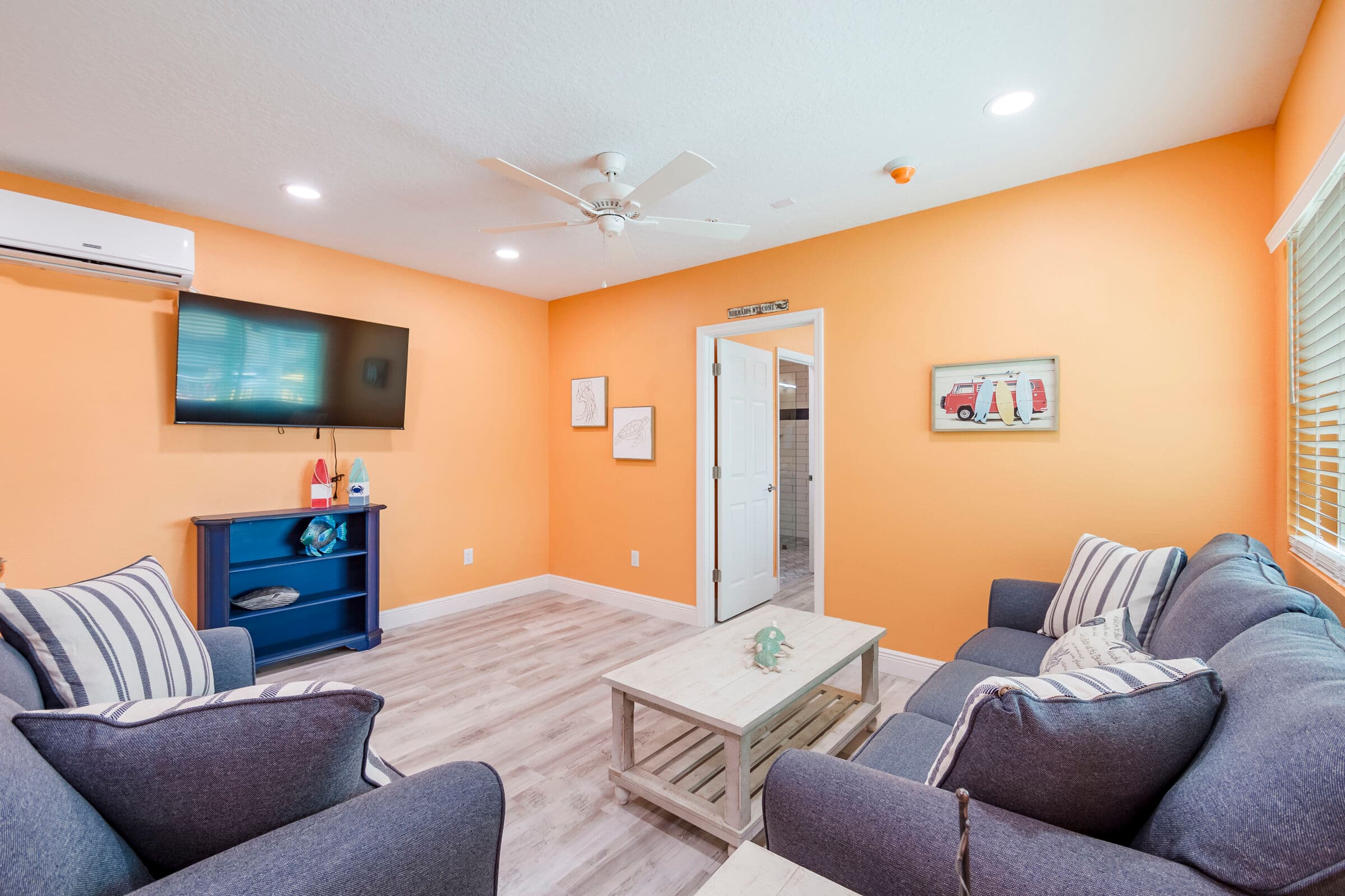A spacious living area and kitchenette featuring orange walls and light wood-style flooring. The room includes a large wall-mounted TV above a blue console table, a grey storage cabinet, and a small black dining set, with a glimpse of a white coffee table and a grey sofa with striped pillows in the foreground.
