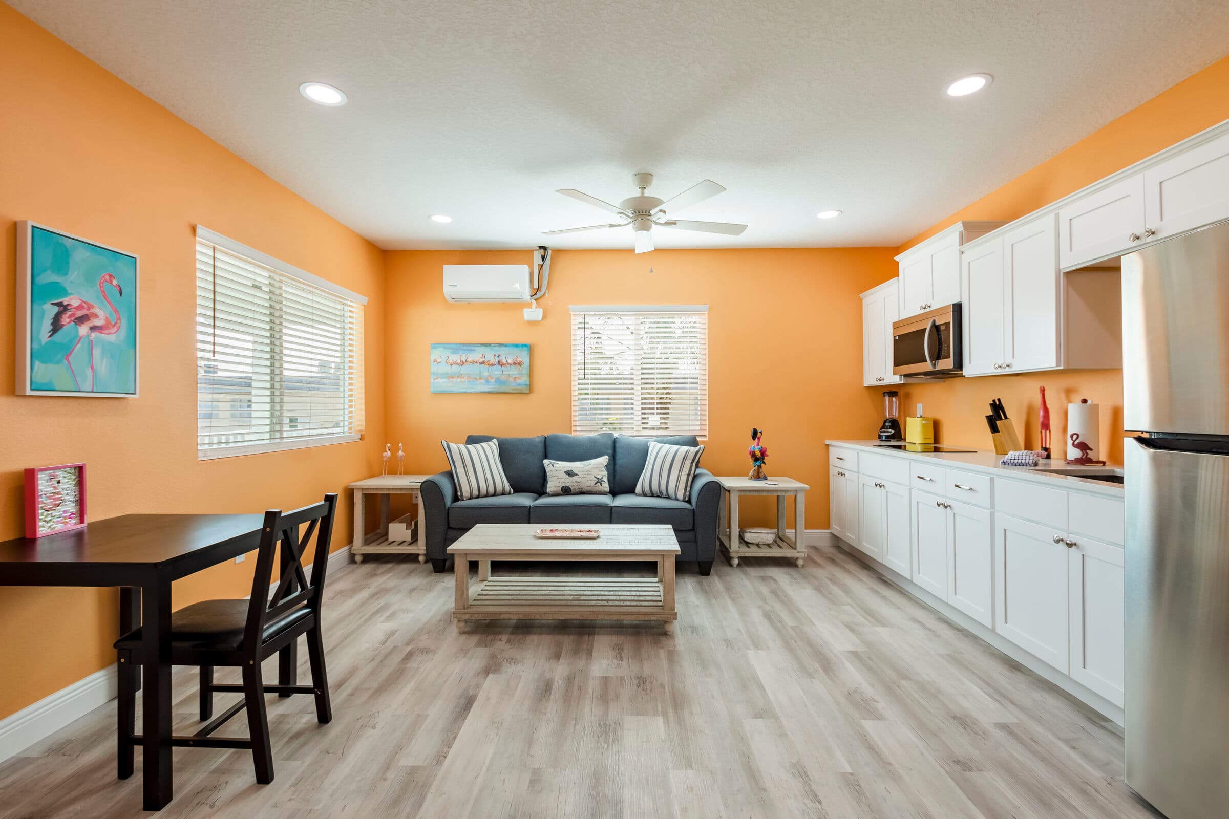 A spacious, open-concept living and dining area with orange walls and light wood-plank flooring. The room features a grey sofa with striped pillows, a white coffee table, a small black dining set, and a full wall of white kitchen cabinetry with stainless steel appliances.