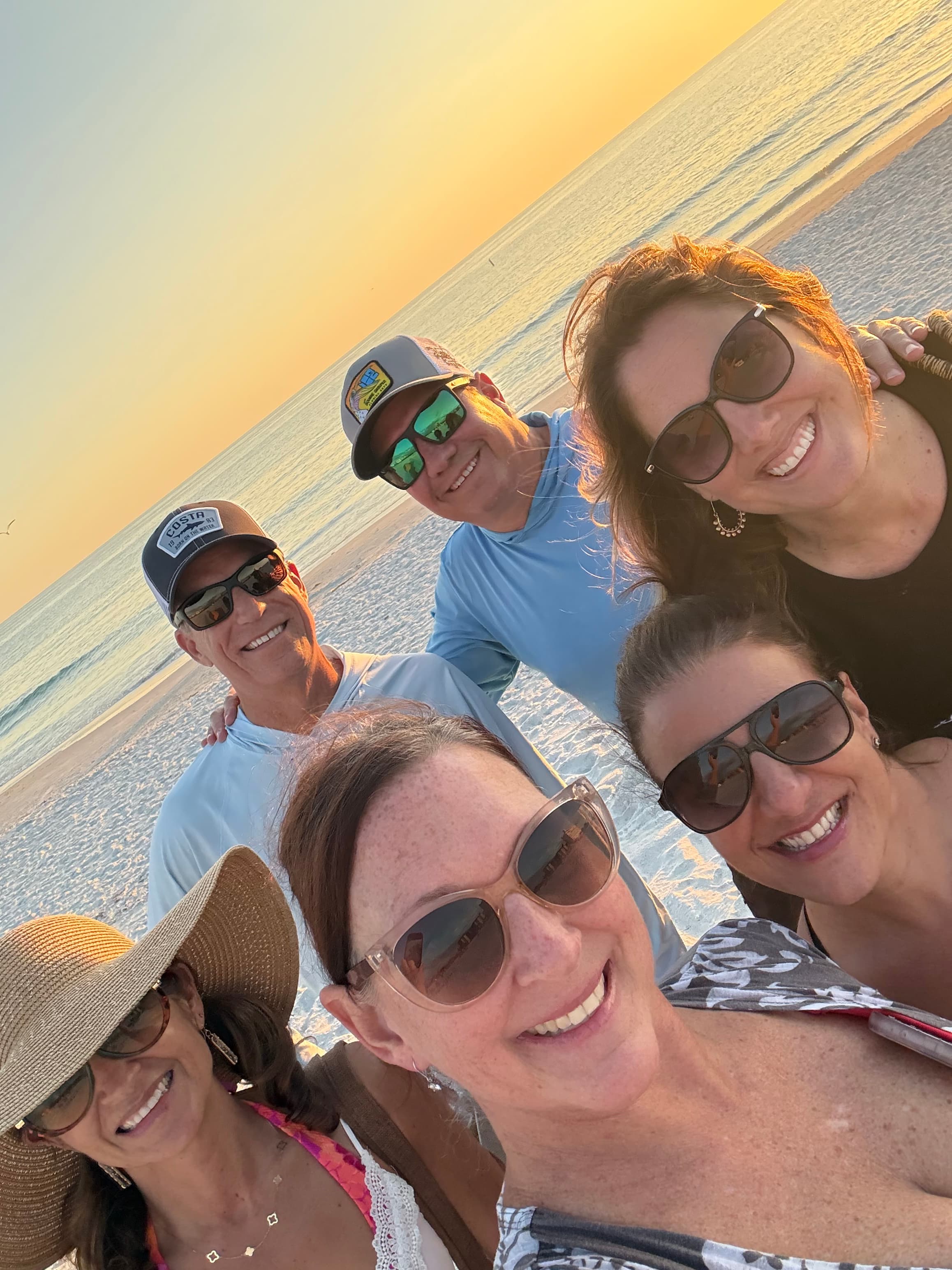 A group of six people smiles for a selfie on the beach during sunset.