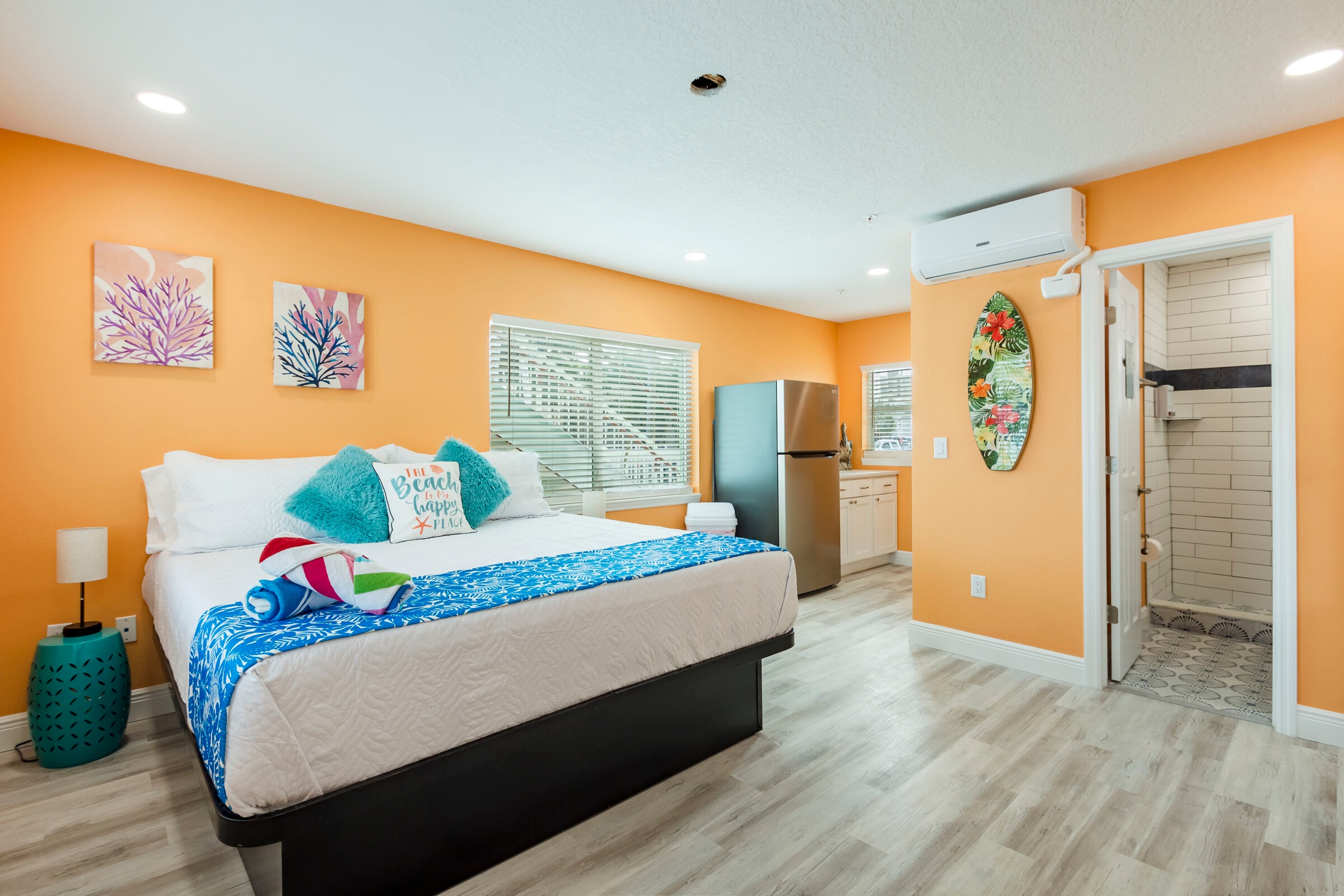 A bright, beach-themed studio accommodation featuring orange walls and light wood flooring. A large bed with white linens and turquoise accent pillows sits in the foreground. In the background, there is a small kitchenette with a stainless steel refrigerator and an open doorway leading to a white-tiled bathroom. A decorative surfboard hangs on the wall near the door.