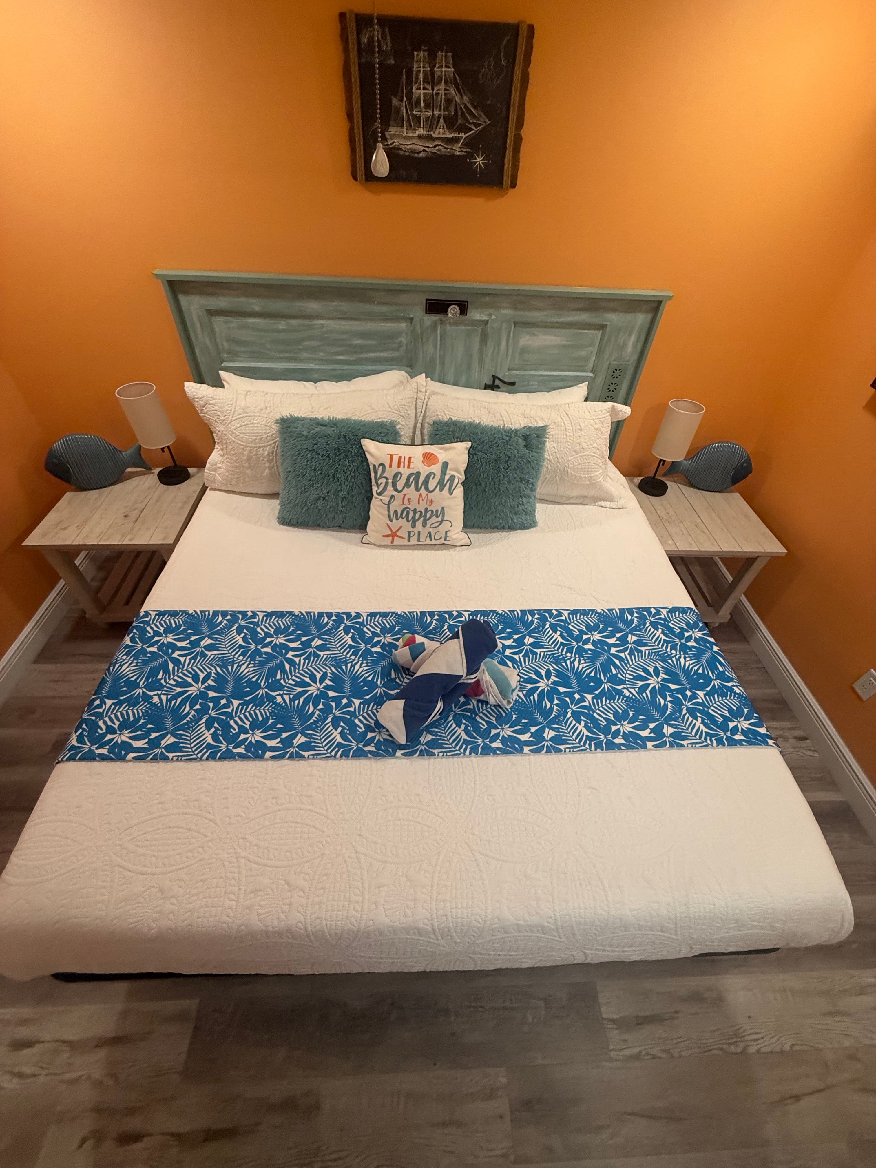 A neatly made bed with teal and white bedding, decorative pillows, and an orange wall backdrop.