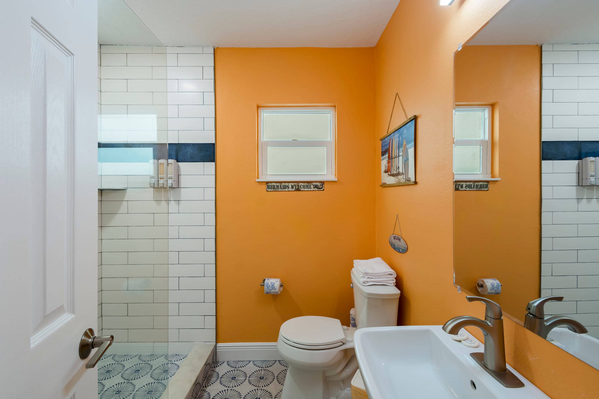 A modern bathroom featuring vibrant orange walls and a white-tiled walk-in shower with a decorative blue mosaic strip. The room includes a white toilet and a white vanity sink with a chrome faucet, positioned beneath a large mirror. Patterned floor tiles in shades of gray and white complete the coastal-inspired look.