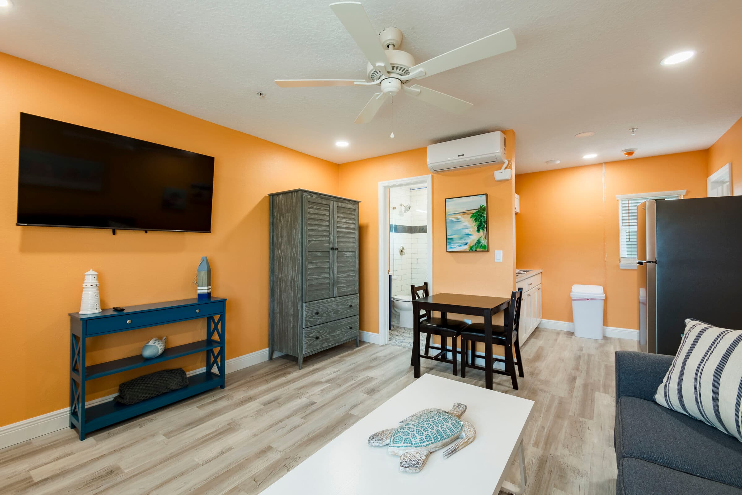 A cozy studio living area with vibrant orange walls and light wood-look flooring. The space features a dark gray sofa with striped accent pillows, a white coffee table with a decorative starfish, and a small black dining table with two chairs. An open doorway on the left leads to a bright white bathroom, and a stainless steel refrigerator is visible in the background.