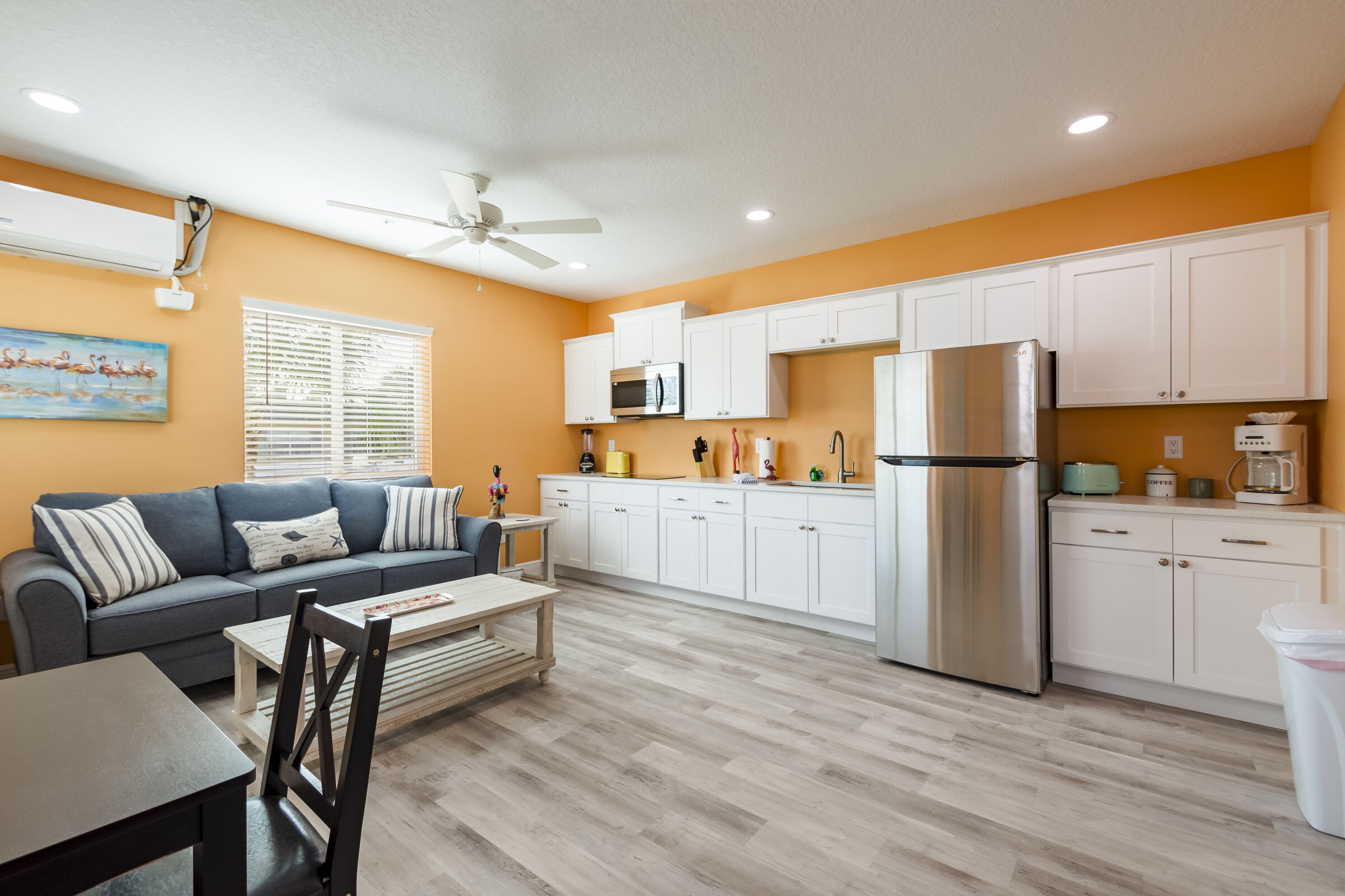 A spacious, open-concept living and dining area featuring orange walls and light wood-style flooring. The space includes a grey sofa with striped pillows, a white coffee table, and a full wall of white kitchen cabinets with stainless steel appliances, including a large refrigerator and a microwave.