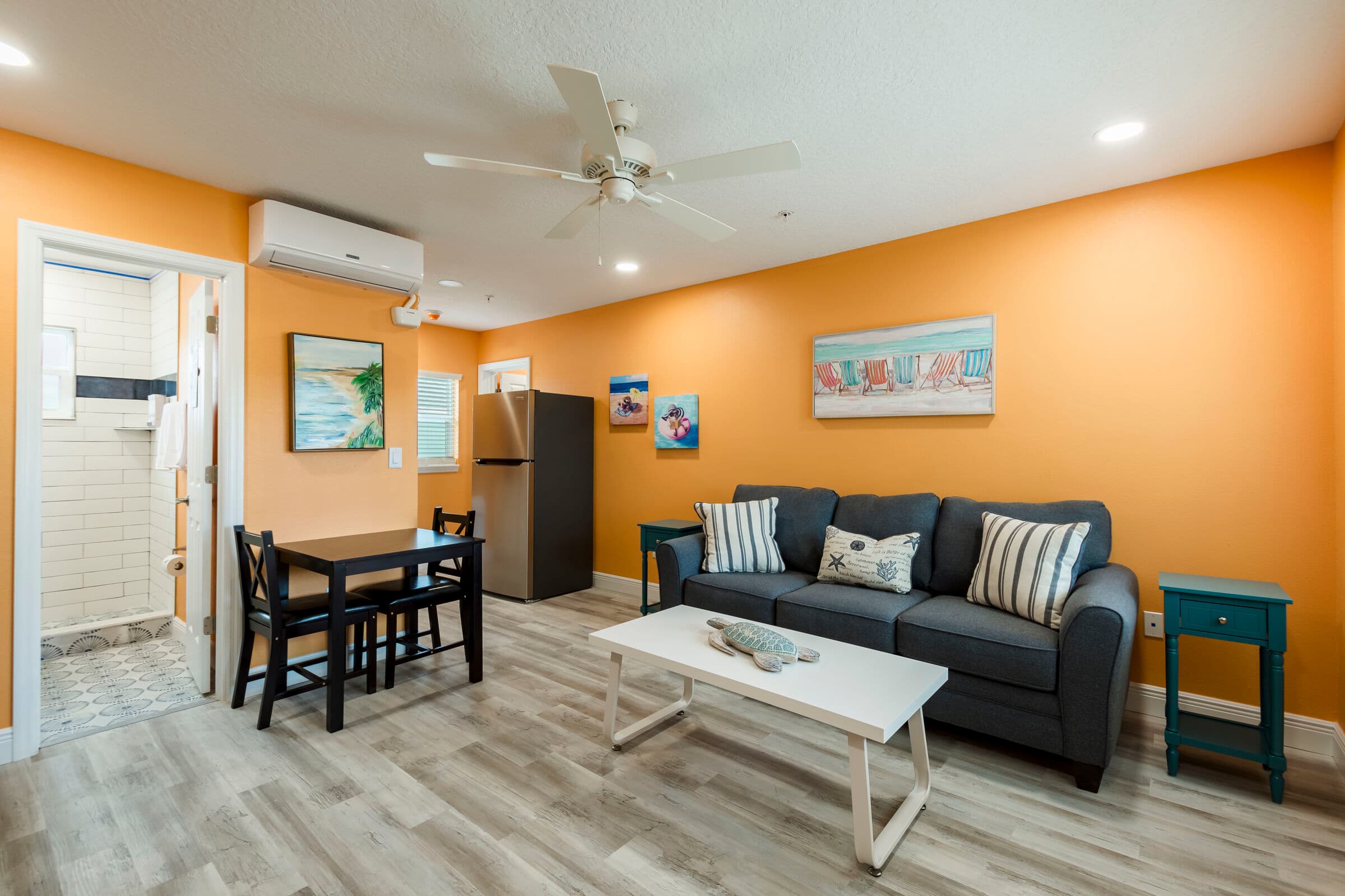 A cozy studio living area with vibrant orange walls and light wood-look flooring. The space features a dark gray sofa with striped accent pillows, a white coffee table with a decorative starfish, and a small black dining table with two chairs. An open doorway on the left leads to a bright white bathroom, and a stainless steel refrigerator is visible in the corner.