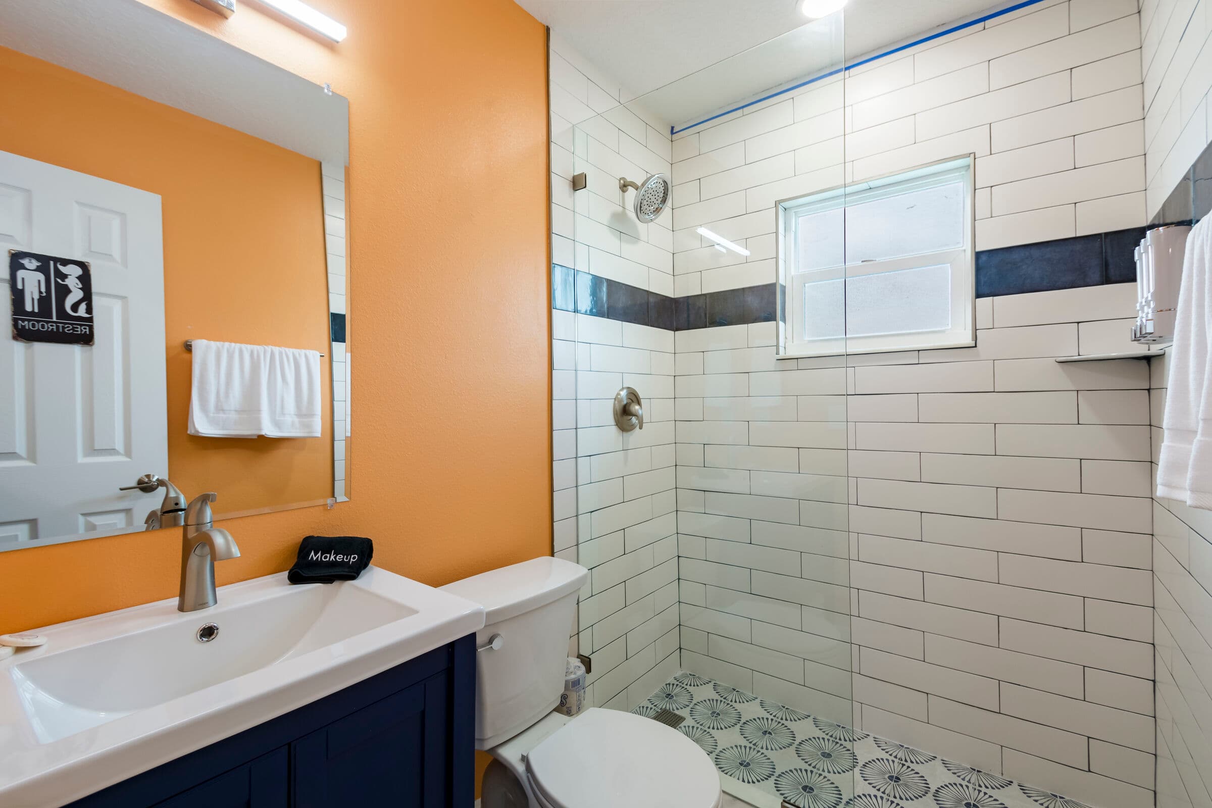 A modern bathroom featuring vibrant orange walls and a white-tiled walk-in shower with a dark blue mosaic accent strip. The room includes a navy blue vanity with a white sink and chrome faucet, a white toilet, and patterned gray and white floor tiles. A large frameless mirror reflects the bright lighting of the space.