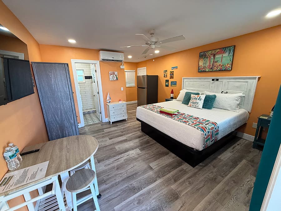 A cozy bedroom featuring an orange accent wall, a queen bed, a small dining table, and a combination of modern and beach-themed decor.