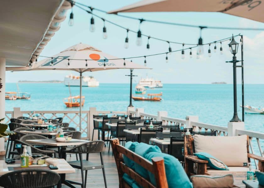 Outdoor waterfront dining area with tables, string lights, and umbrellas overlooking boats on the water.