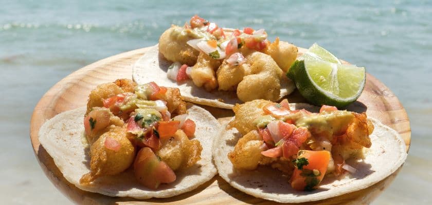 Three fish tacos topped with pico de gallo and sauce served on a wooden board with a lime wedge by the beach.