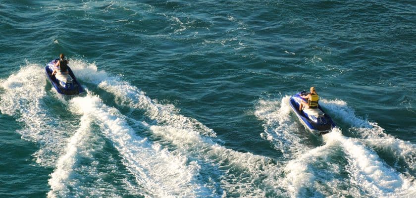 Two people ride jet skis across the ocean, leaving white wakes behind them on the water.