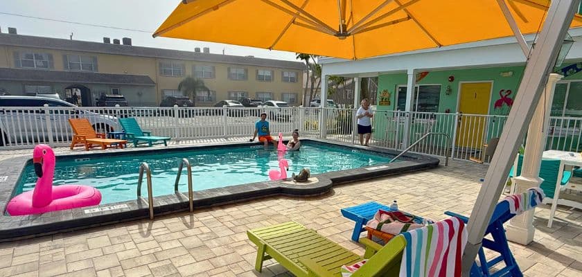 The pool at The Lofts features lounge chairs, flamingo floats, and guests enjoying a sunny afternoon beside the water.