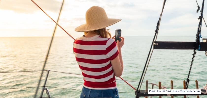 A woman wearing a sun hat and striped shirt takes a photo while standing aboard a sailboat during golden hour on the water.